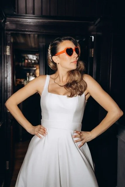 Bride standing confidently in a short white reception dress with her hands on her hips, wearing heart-shaped sunglasses