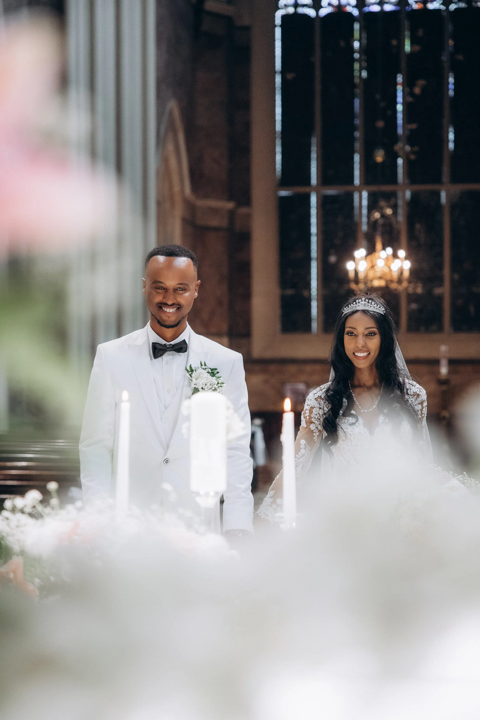 Atmospheric shot of the bride and groom at the altar framed by white candles and soft floral arrangements during a romantic London church wedding.