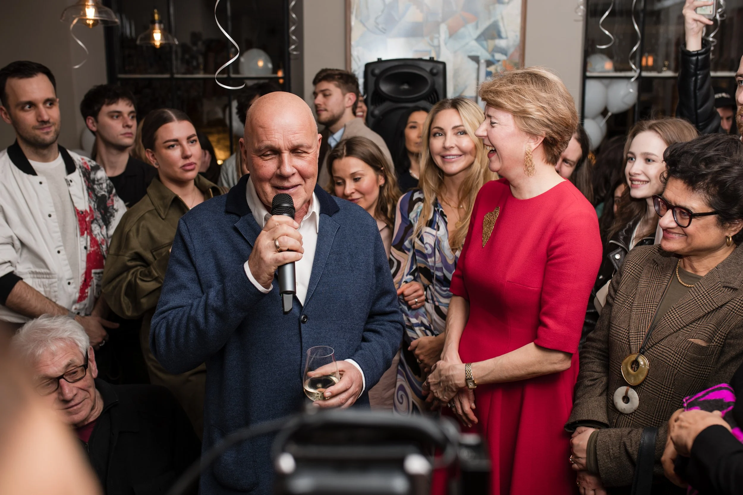A man in a blue blazer speaking into a microphone at a gathering, with a woman in a red dress smiling beside him, and a group of people listening and socializing in the background.
