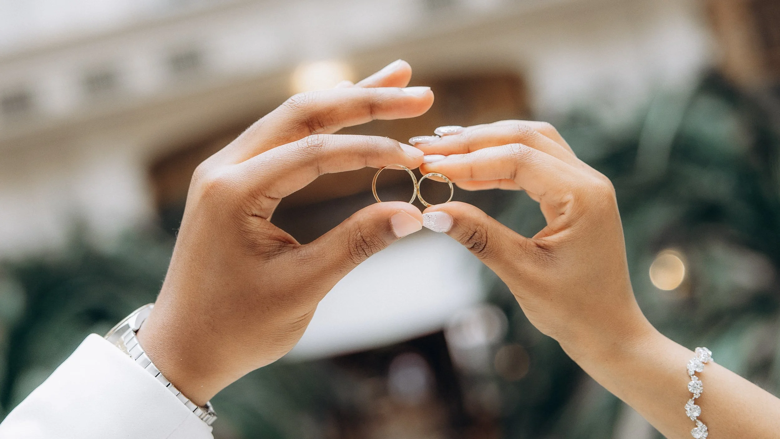 A creative macro/close-up detail shot of the couple’s two gold wedding bands held between their fingertips, with a very blurred, soft background, focusing on the symbolism of commitment and partnership.