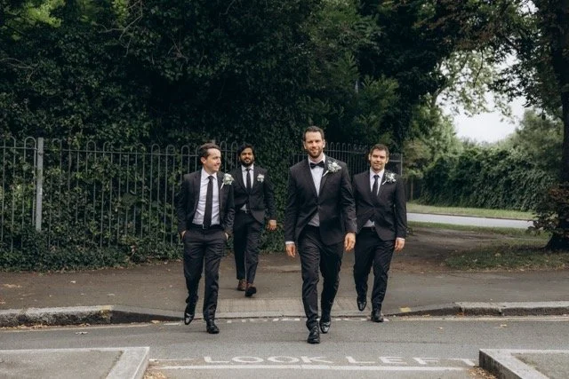 Three groomsmen in black suits walking side by side on a paved road