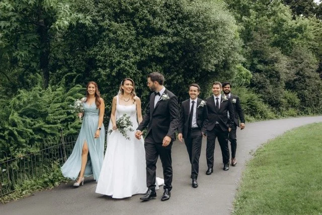 Bridal party walking along a garden path — two bridesmaids in sage green and the couple