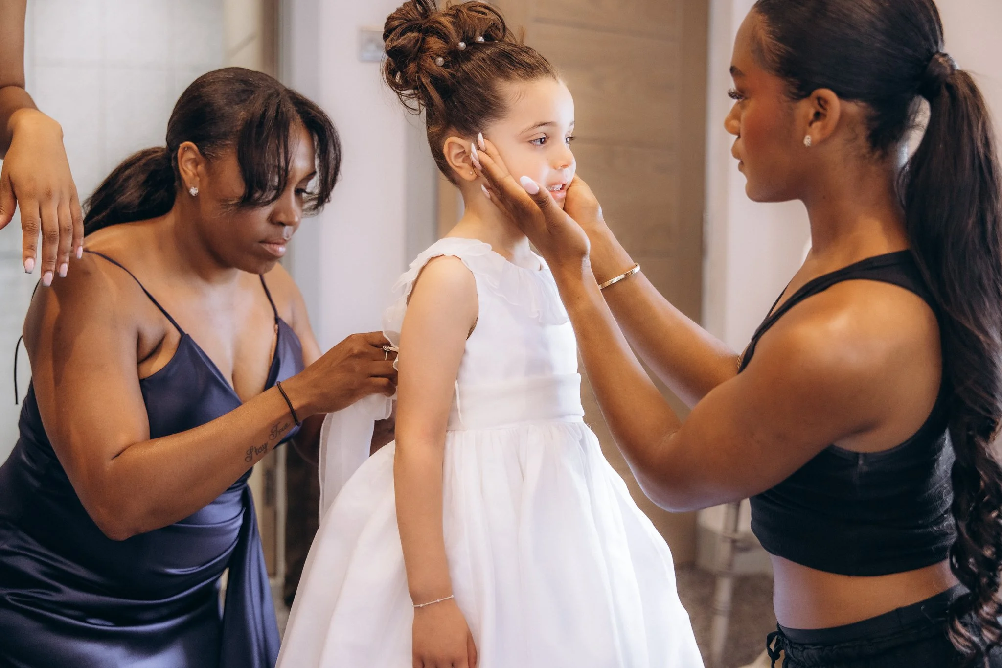 Bridesmaid applying makeup to a young flower girl in a white dress