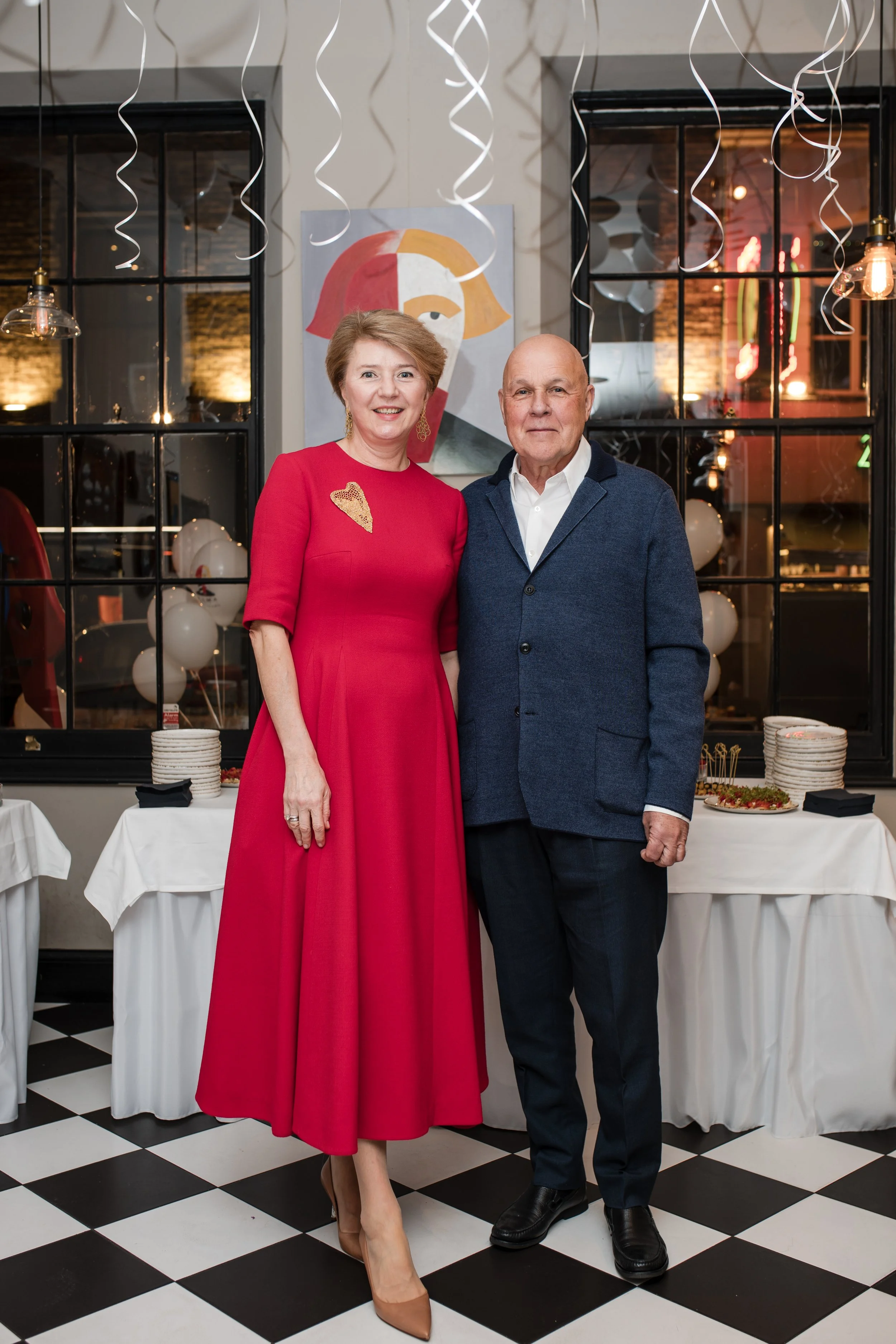 A woman wearing a red dress and beige heels standing beside a man in a dark blazer and white shirt. They are in a decorated space with black-and-white checkered floor, white balloons, and streamers, and a colorful abstract portrait on the wall behind them.