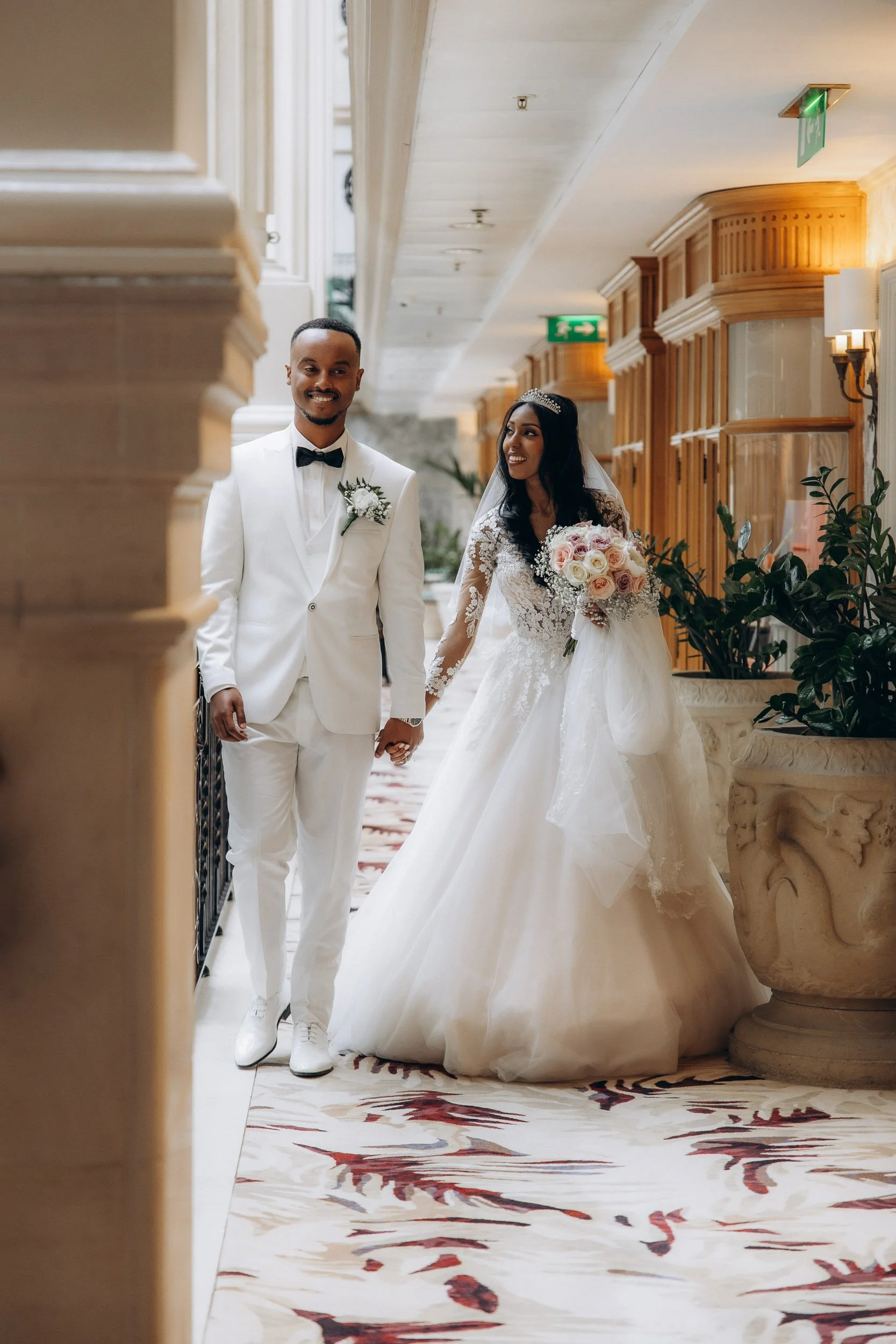 Recessional walk down the grand hallway of The Landmark Hotel, as the newly married couple, bride in a full gown and groom in all-white, hold hands with warm smiles, demonstrating a natural, joyful documentary photography style.