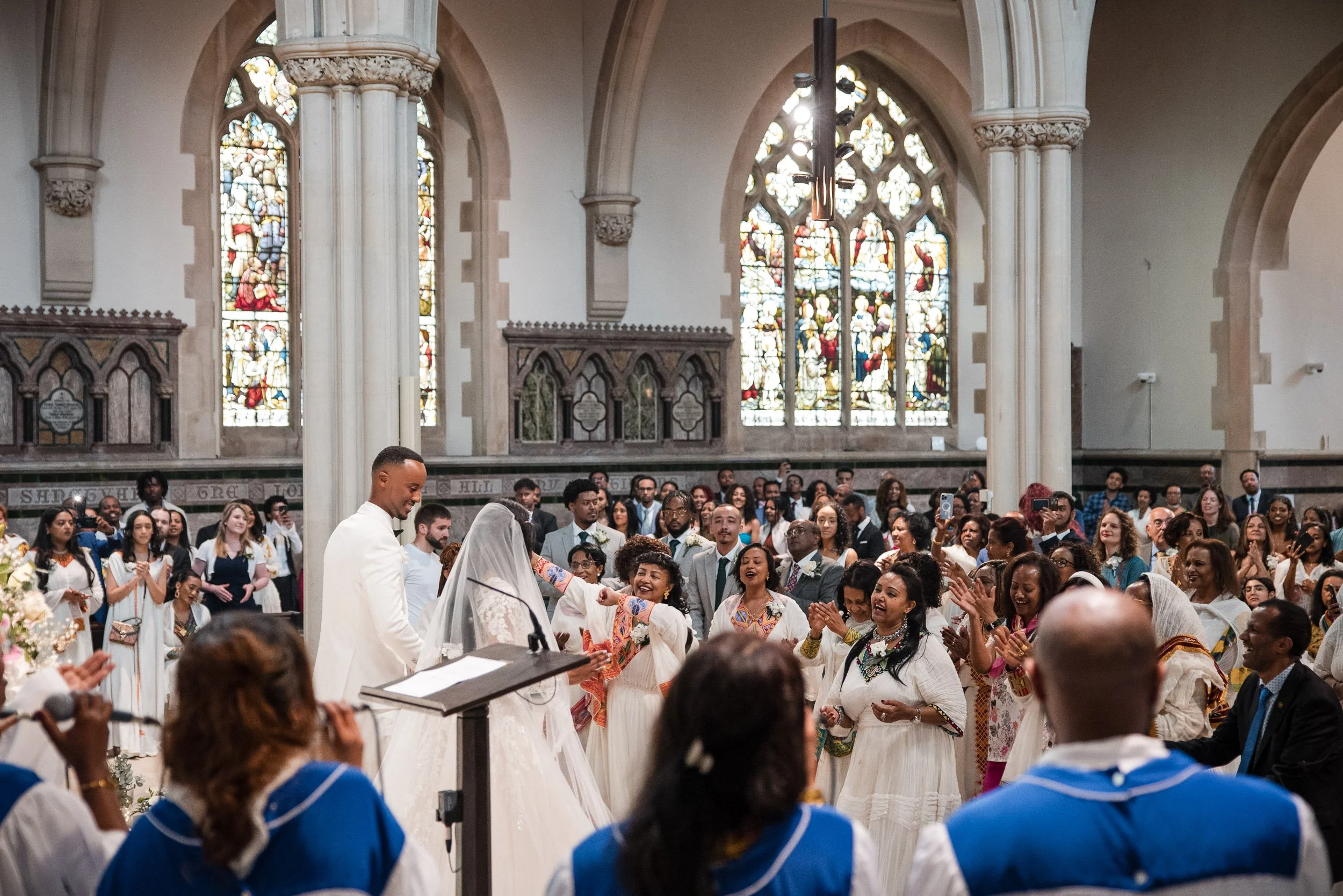 Perspective shot of a wedding ceremony in a historic London church with large stained glass windows, showing the couple at the altar surrounded by their wedding guests.