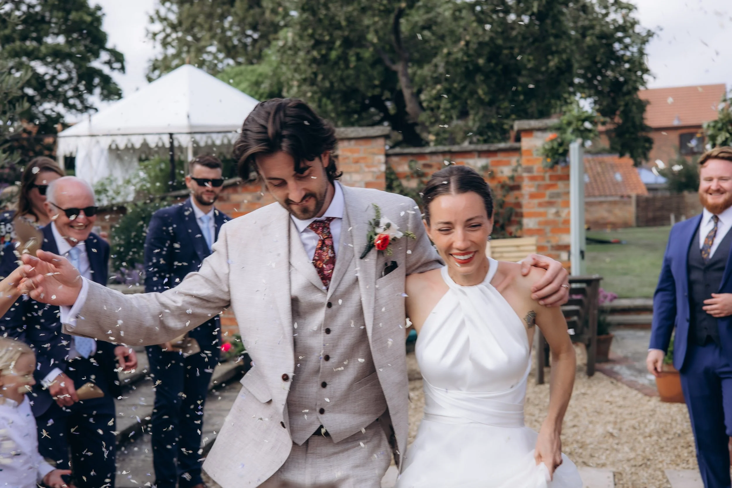 Bride and groom walking arm in arm through a confetti shower past a stone-walled farmhouse decorated with red and pink geraniums, the groom grinning widely as confetti rains down