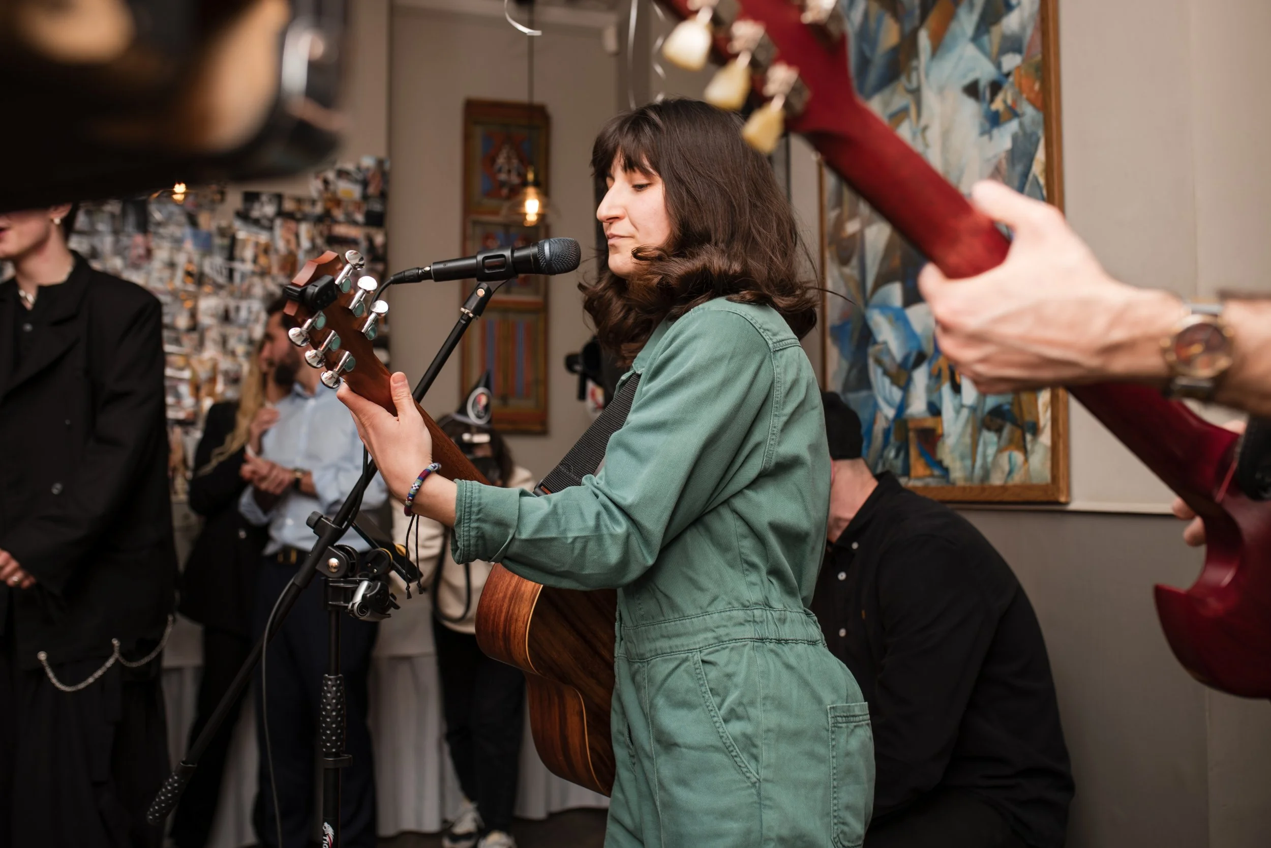 A woman with dark brown hair in a green jacket playing an acoustic guitar and singing into a microphone during a live performance or jam session in an art gallery or similar indoor setting.