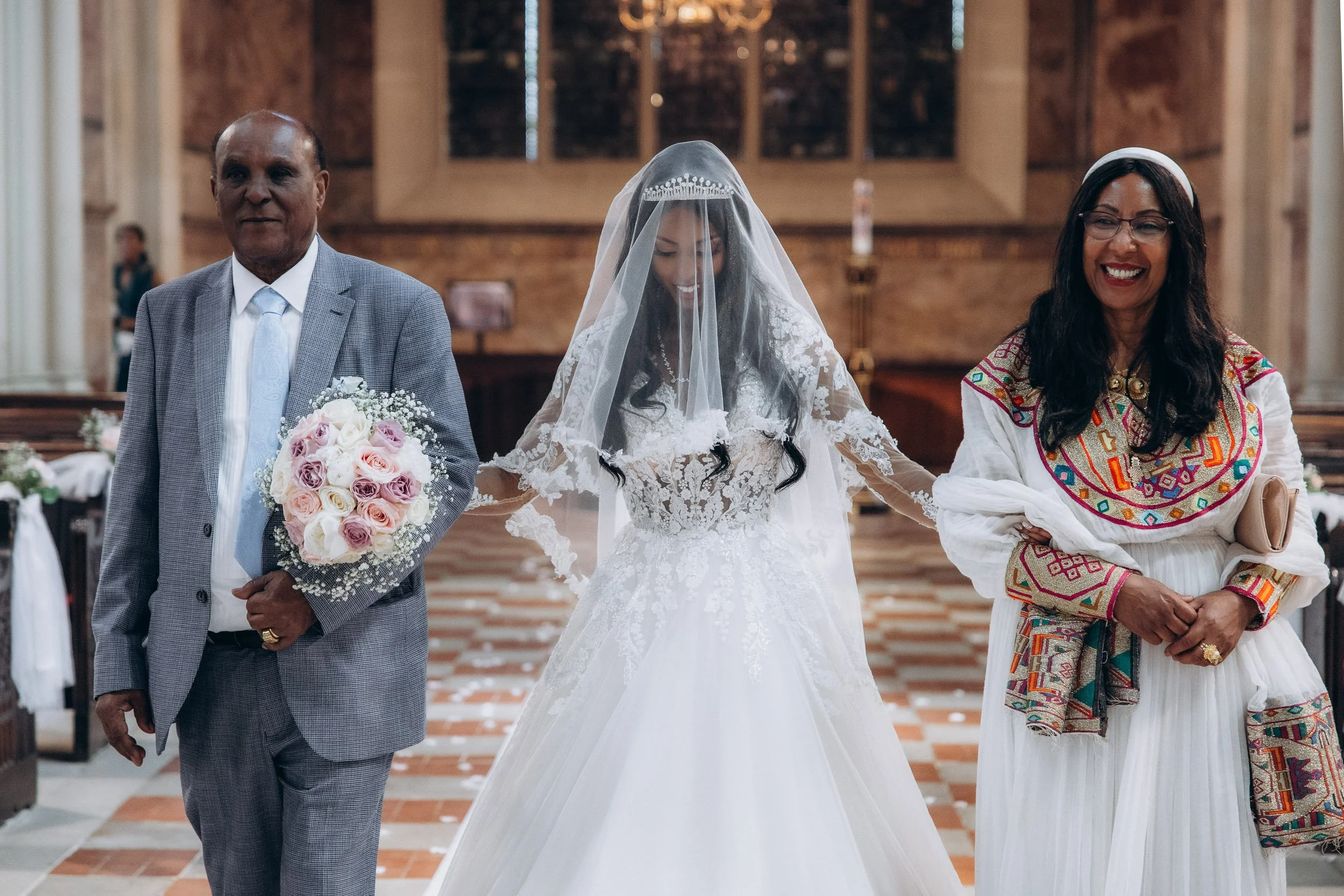 A stunning bride in a lace wedding gown and long veil walking down the aisle of a candlelit London church, escorted by her father.