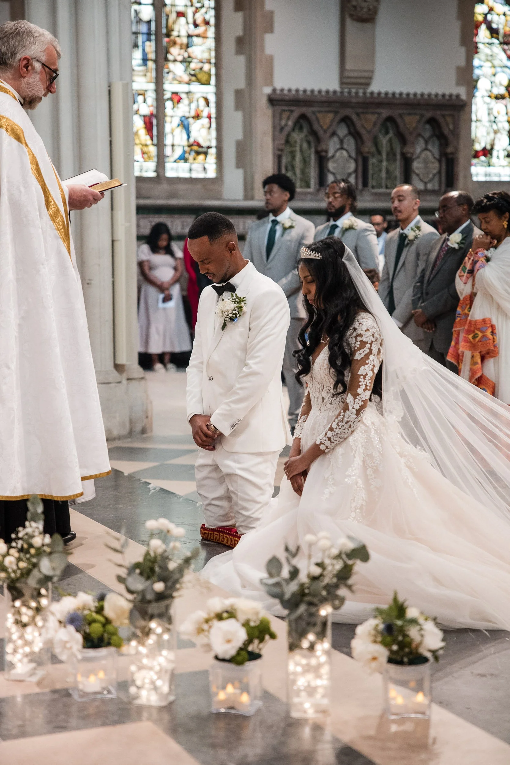A deeply spiritual moment as the bride and groom kneel on colorful cushions during their Orthodox wedding ceremony, receiving a blessing from a priest (out of frame) in a historic London church with a large stained-glass window in the background.