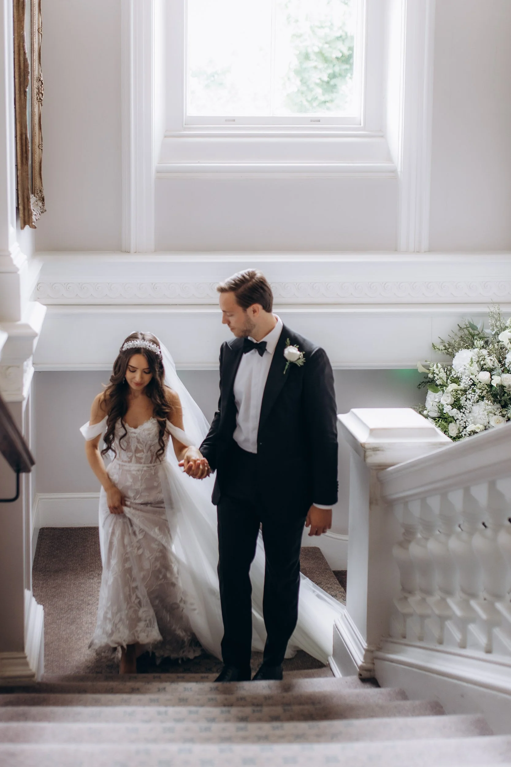Elegant bride and groom in a black tuxedo walking down a grand white staircase at a luxury London wedding venue; sophisticated and classic wedding portraits.