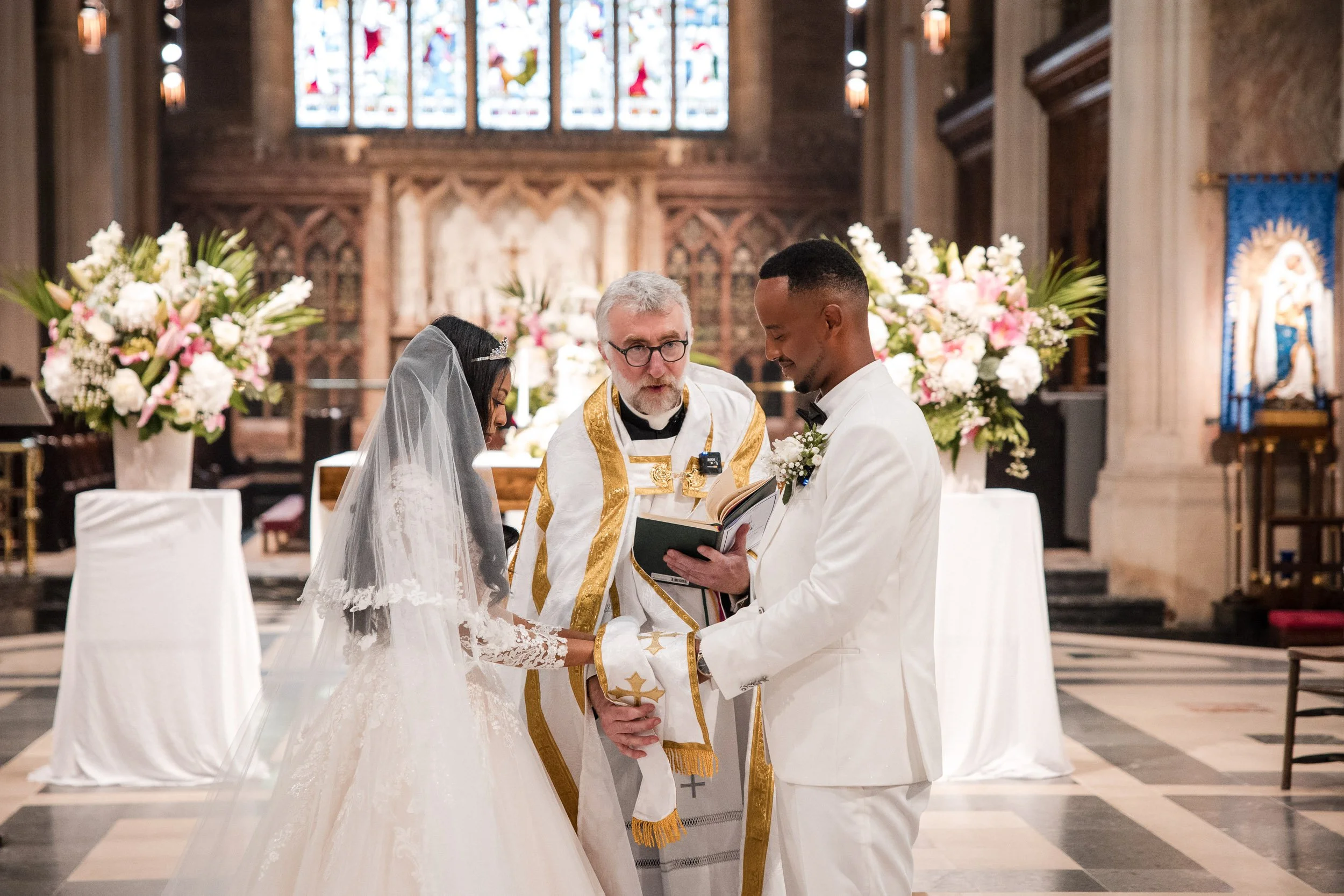 Close-up of an elderly Ethiopian Orthodox priest in traditional red and gold vestments blessing the bride and groom during their wedding vows in a London church, emphasizing the sacred and traditional nature of the ceremony.