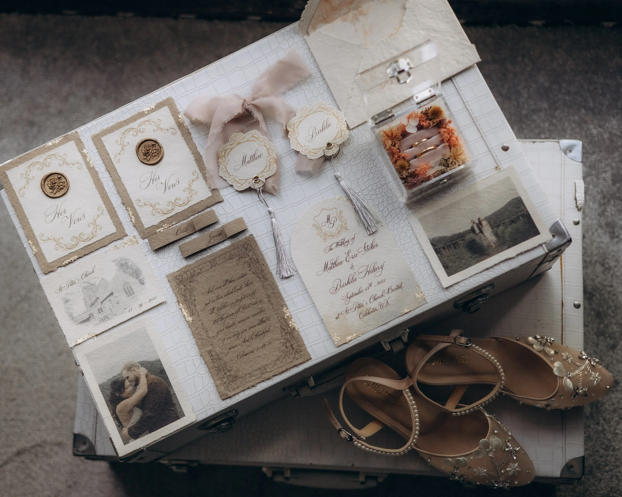 A display of wedding invitations, name tags, and jewelry on a white textured surface. The invitations feature elegant script with gold accents, and the jewelry includes rings and a pair of nude heels with pearl and floral embellishments.