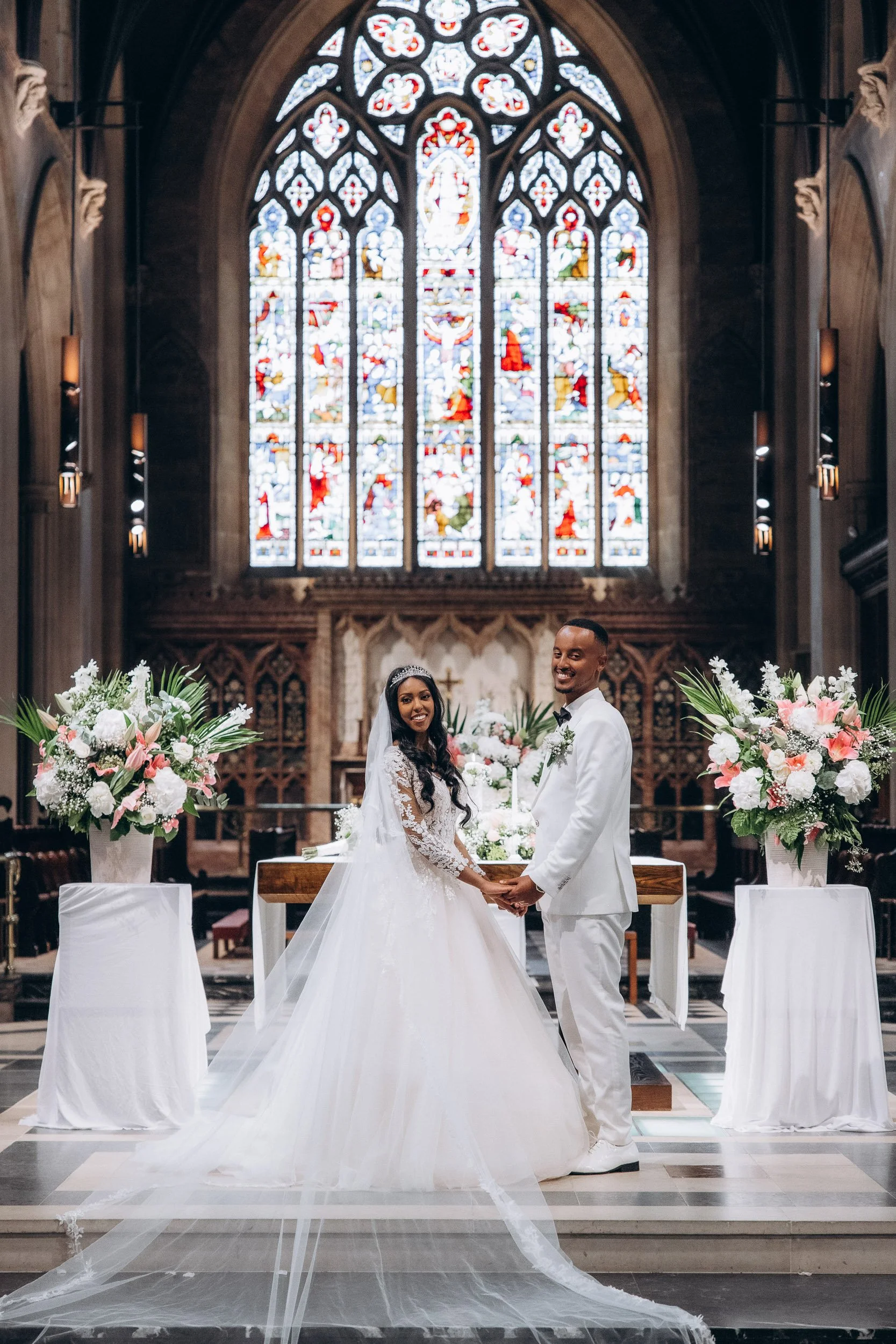 Wide-angle portrait of the bride and groom holding hands at the altar under a magnificent stained glass window at a luxury London wedding venue.