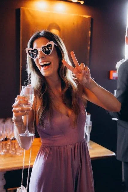 Guest in a short dress wearing heart-shaped novelty sunglasses and holding a champagne flute