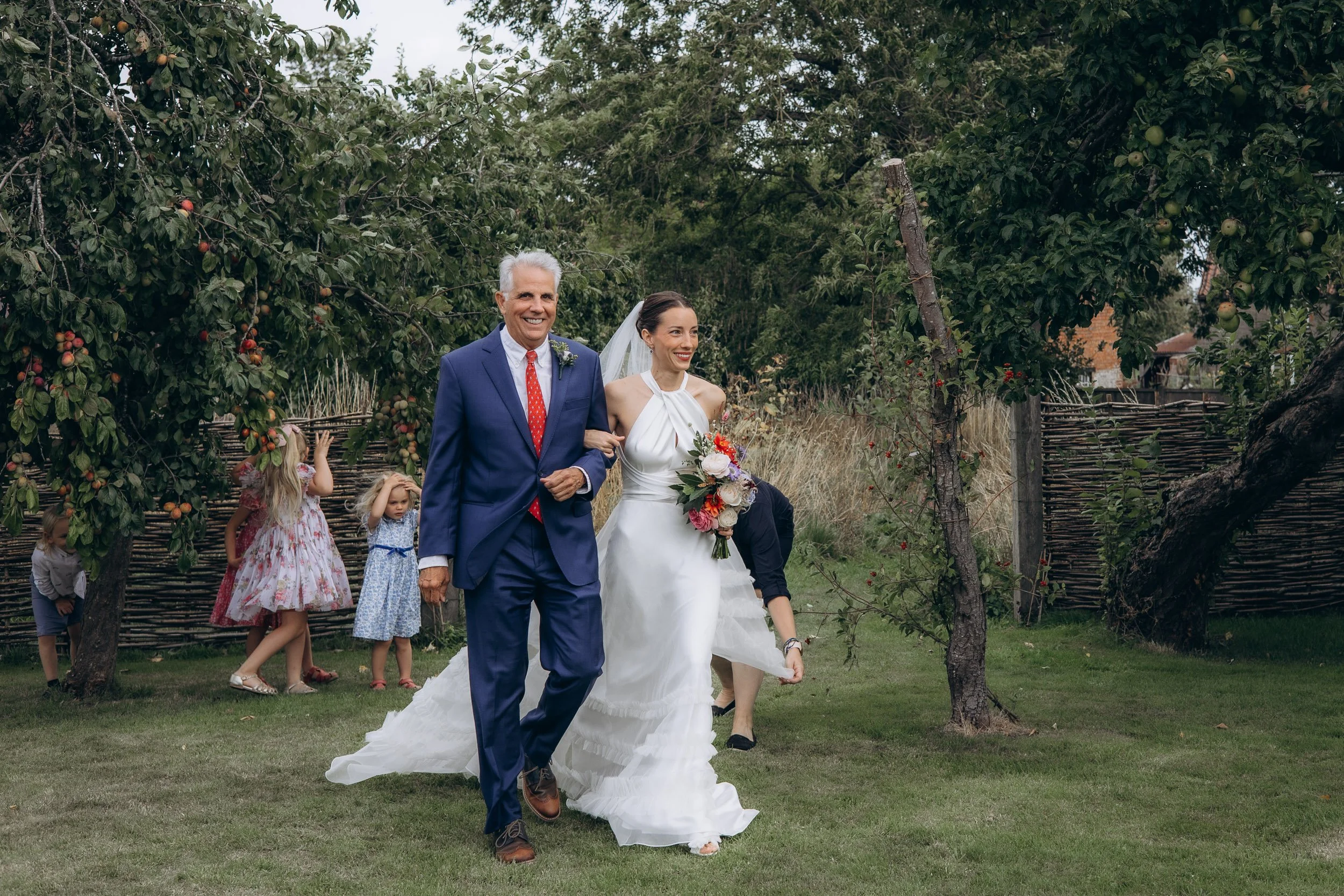 Bride in a white halterneck gown and veil walking arm in arm with her father in a navy suit and red tie through an orchard, both smiling, with children playing in the background