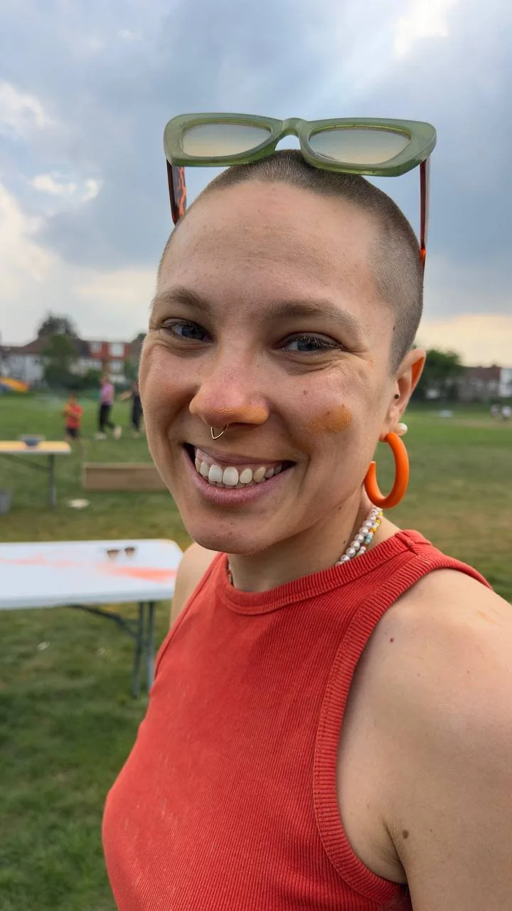 A smiling person with short hair, wearing a red sleeveless top, orange hoop earrings, a beaded necklace, and sunglasses on top of their head standing outdoors in a park.