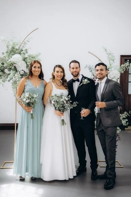 Group of three — bride, groom, and a guest — posing and smiling in front of the floral arch