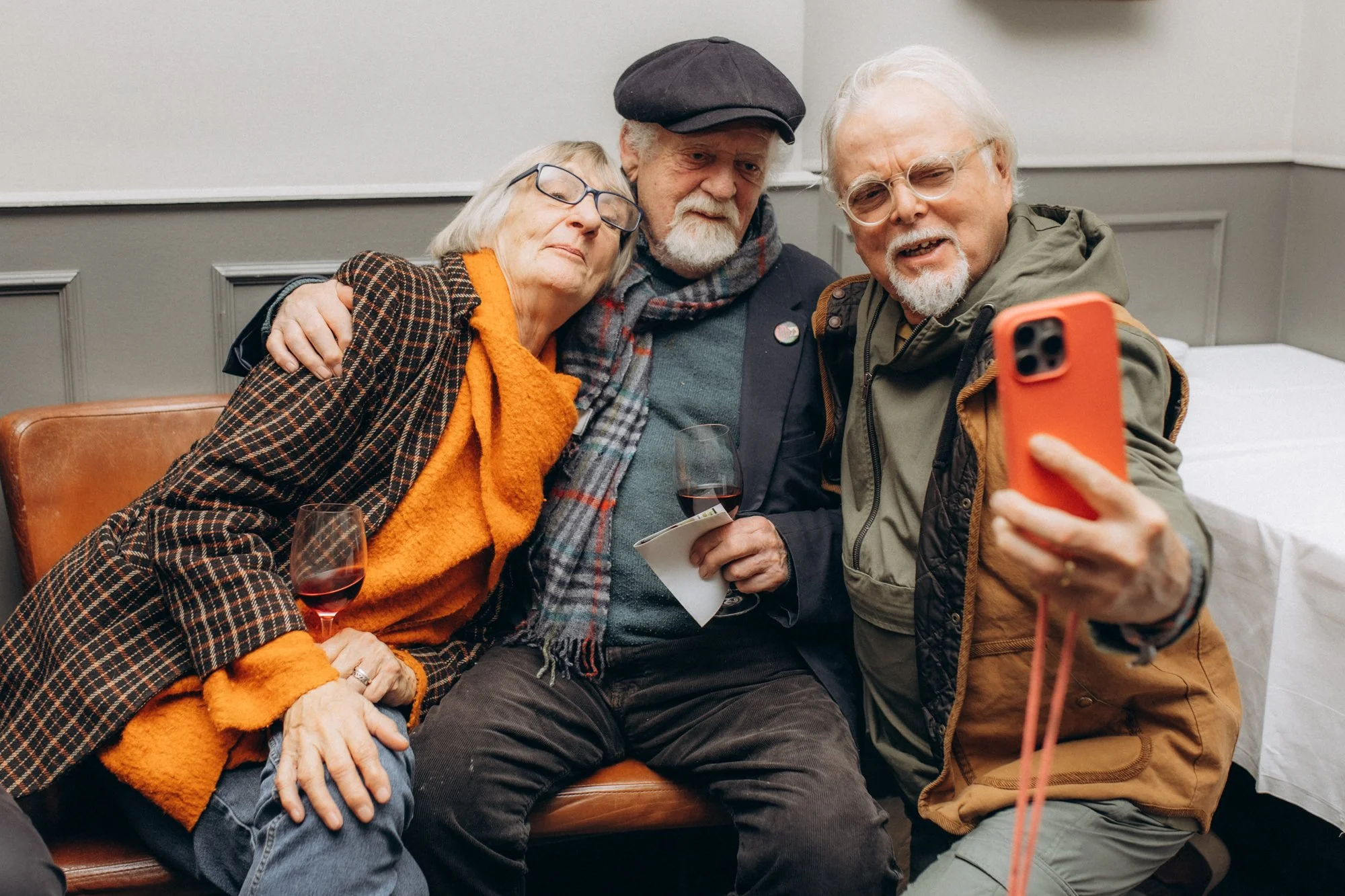 Three elderly people sitting closely together on a bench, taking a selfie with a smartphone. They are in a cozy indoor setting, with two women and one man, all wearing casual warm clothing and glasses. They appear happy and relaxed.