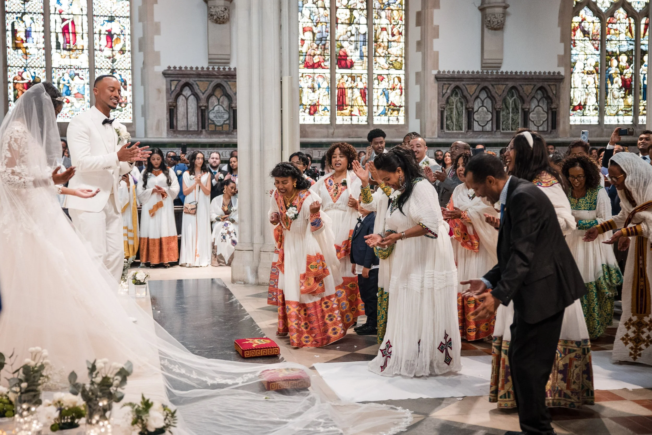 Wide-angle environmental photograph from the aisle of a historic London church, capturing the final blessing of the wedding, showing the couple, the priests, the ornate stained-glass windows, and the entire congregation gathered for the luxury celebr