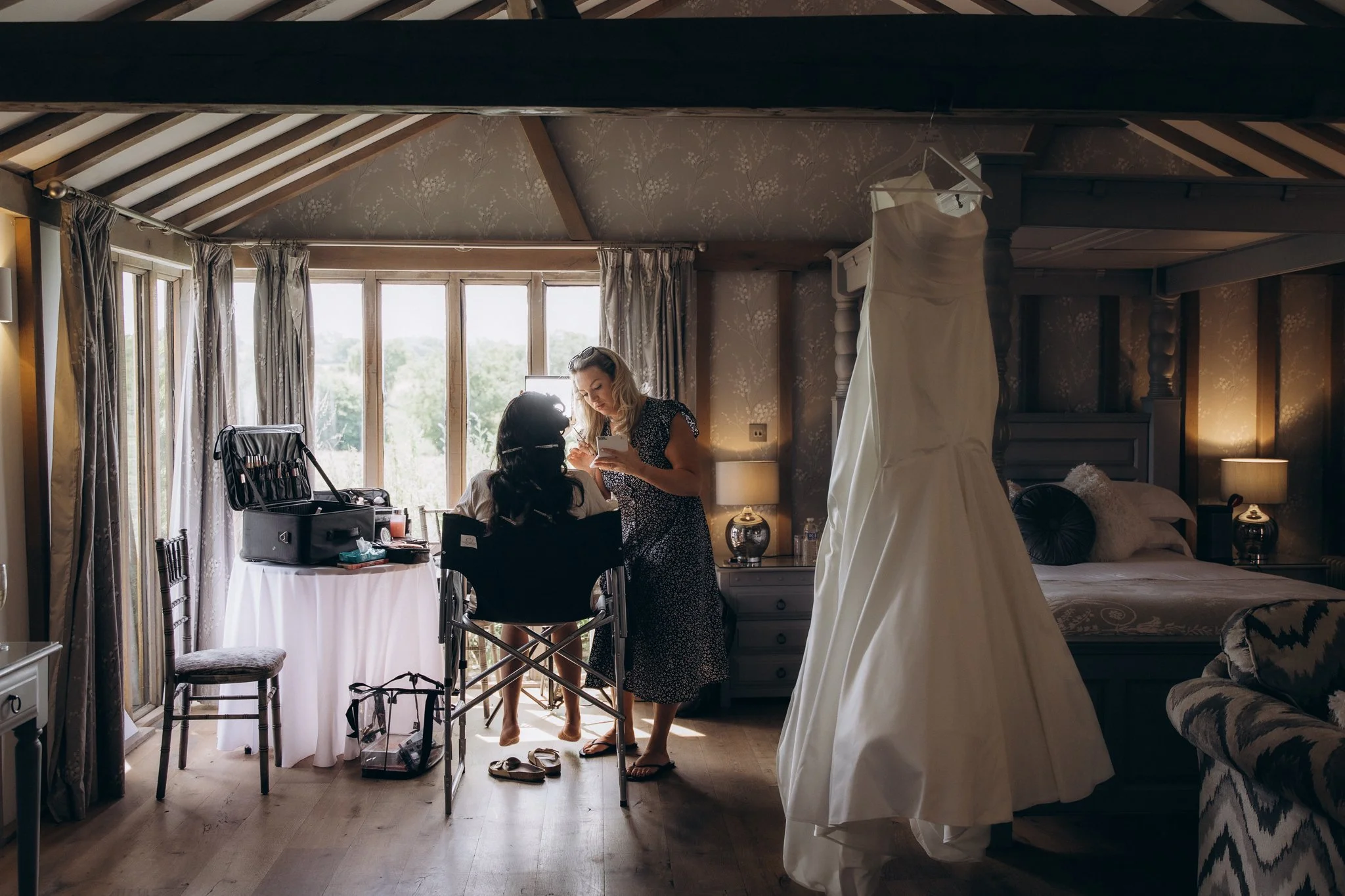 Wedding dress hanging by a window in a rustic barn room as the bride has her hair styled in the background