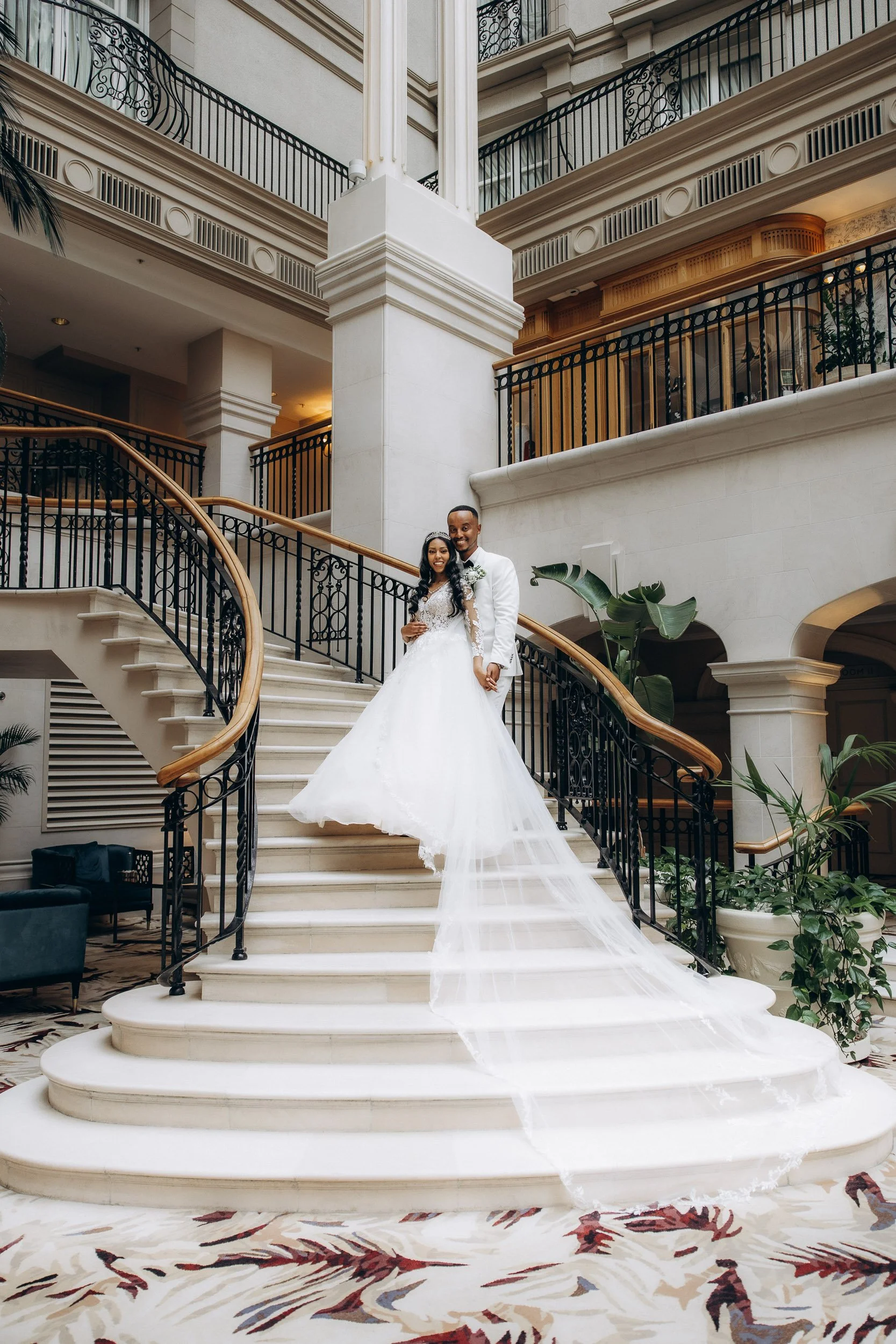 Elegant bride and groom posing on a grand marble staircase at a luxury London hotel wedding venue; high-end London wedding photography by Lusy Klintsova.