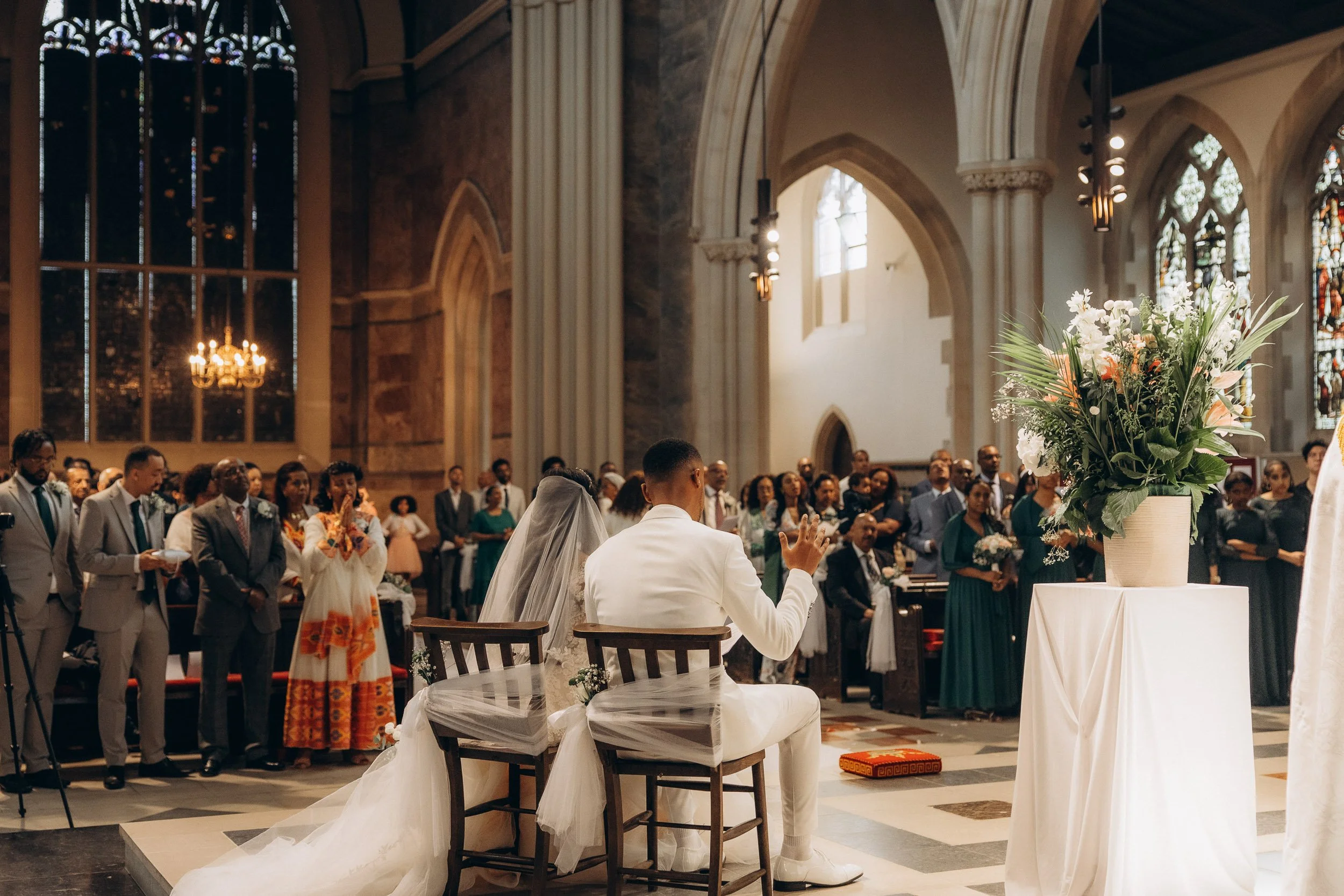 A detailed documentary shot of a traditional Ethiopian Orthodox wedding ceremony, showing the priest in green and the congregation from behind, capturing the atmosphere, the stained glass window, and the sanctity of the church venue in London.