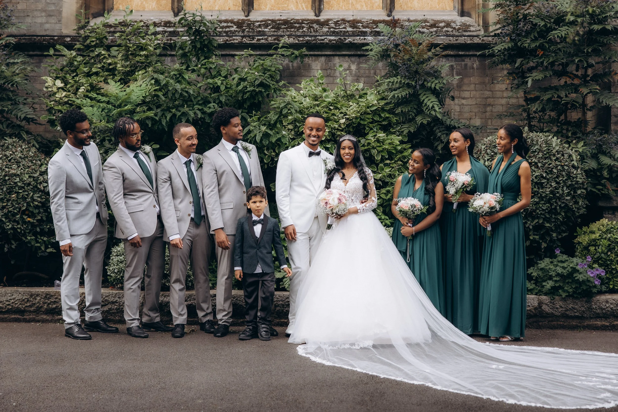 Full-length portrait of a bride and groom with their bridesmaids in forest green dresses and groomsmen in grey suits outside a historic stone church in London.
