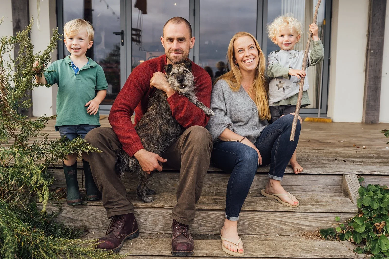 A family of four sitting on wooden steps outside their house, with two young children, a man, and a woman, accompanied by a dog. The children are smiling, and the parents are smiling as well.