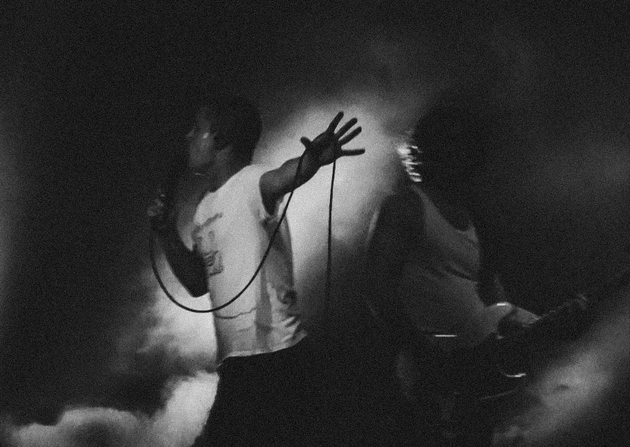 A black and white photo of a man singing into a microphone, with his hand raised, against a smoky background.