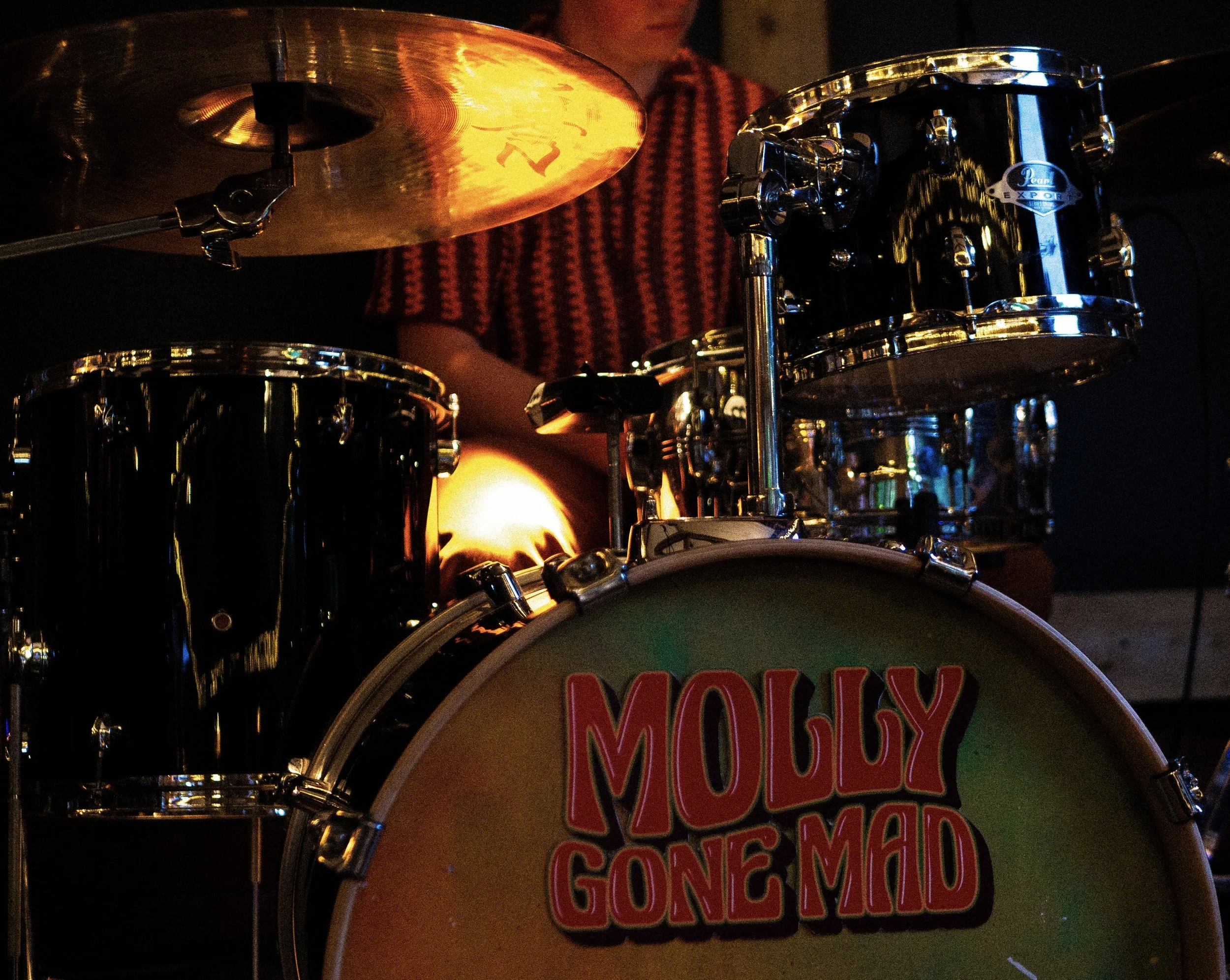 Close-up of a drum set with a sticker reading 'MOLLY GONE MAD', a cymbal, and a person in a red and black checkered shirt in the background at a music performance or rehearsal.