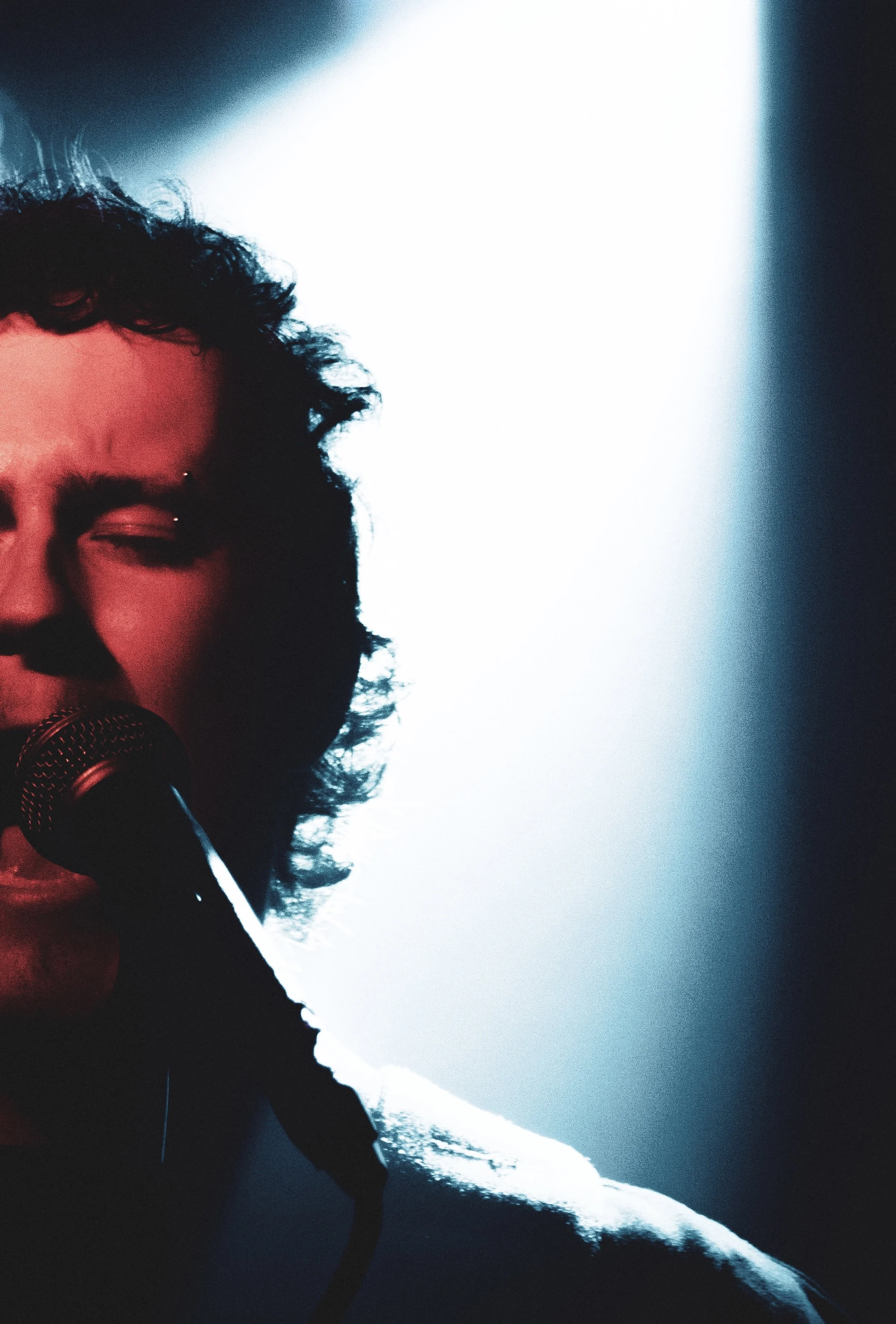 Partially visible man with curly hair singing into a microphone, with a bright light behind him.