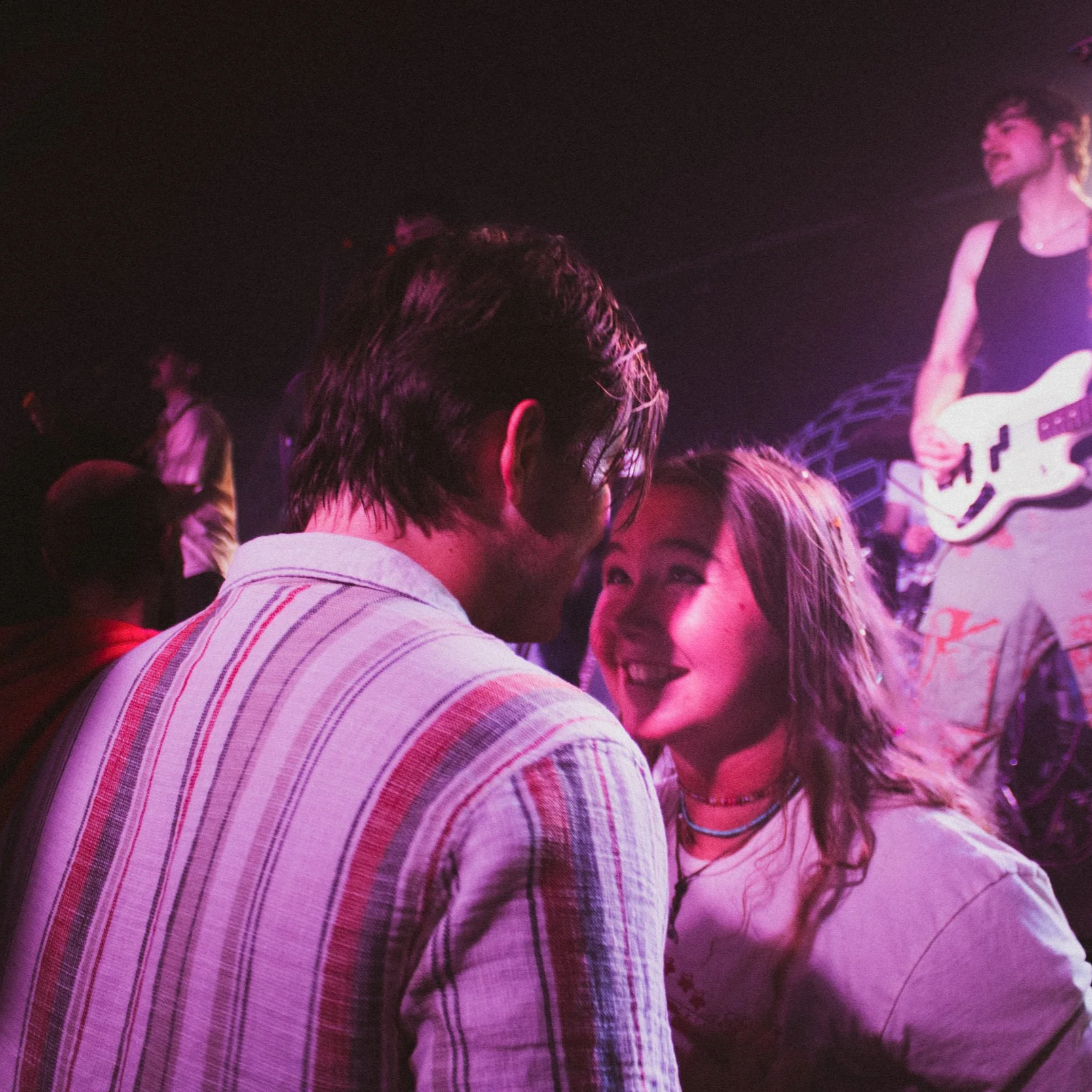 A couple smiling and looking into each other's eyes at a concert or music venue with a musician playing guitar in the background.
