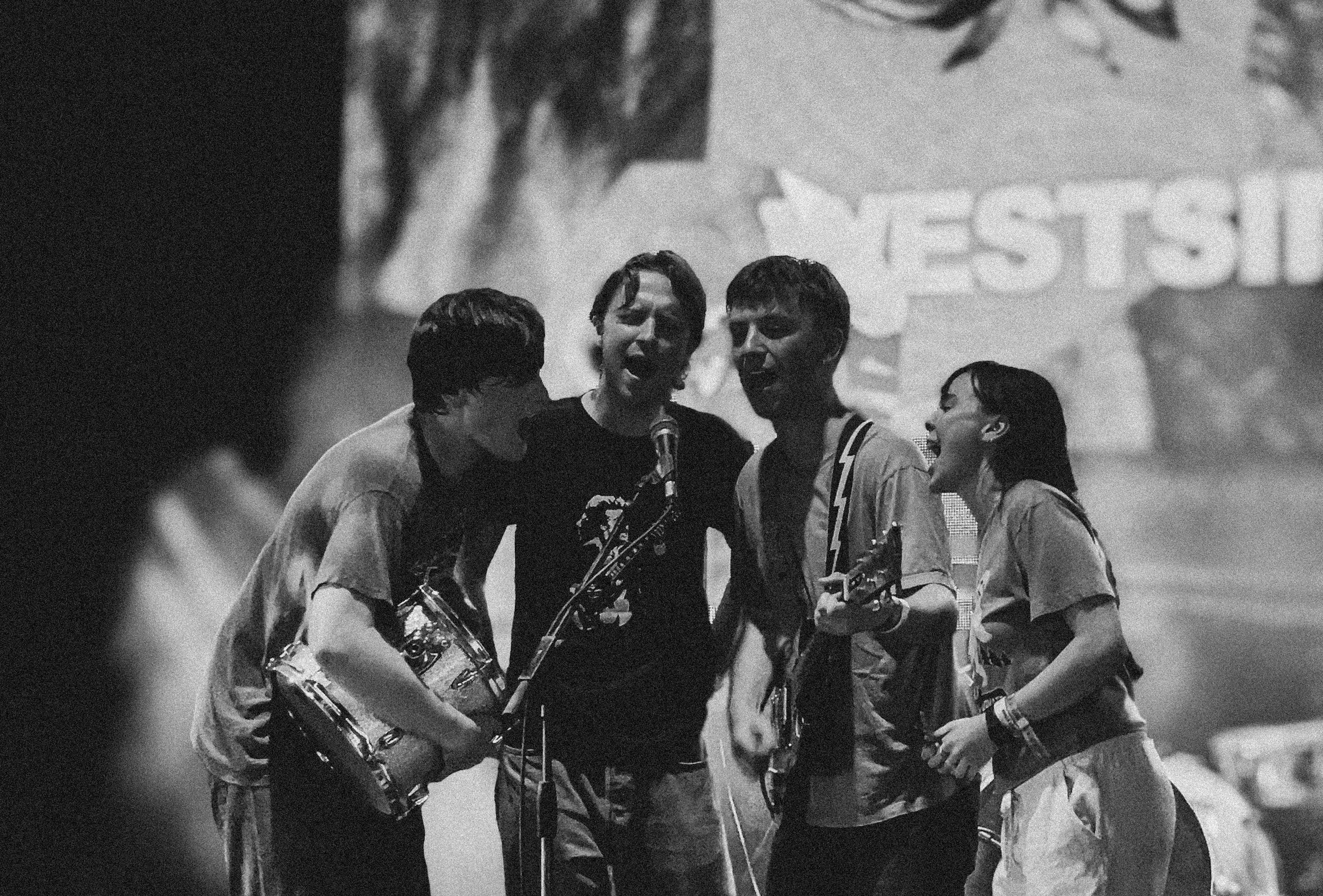 Black and white photo of four young people singing and playing guitars on stage with a large screen behind them that displays the word Westside Cowboy at Truck Festival