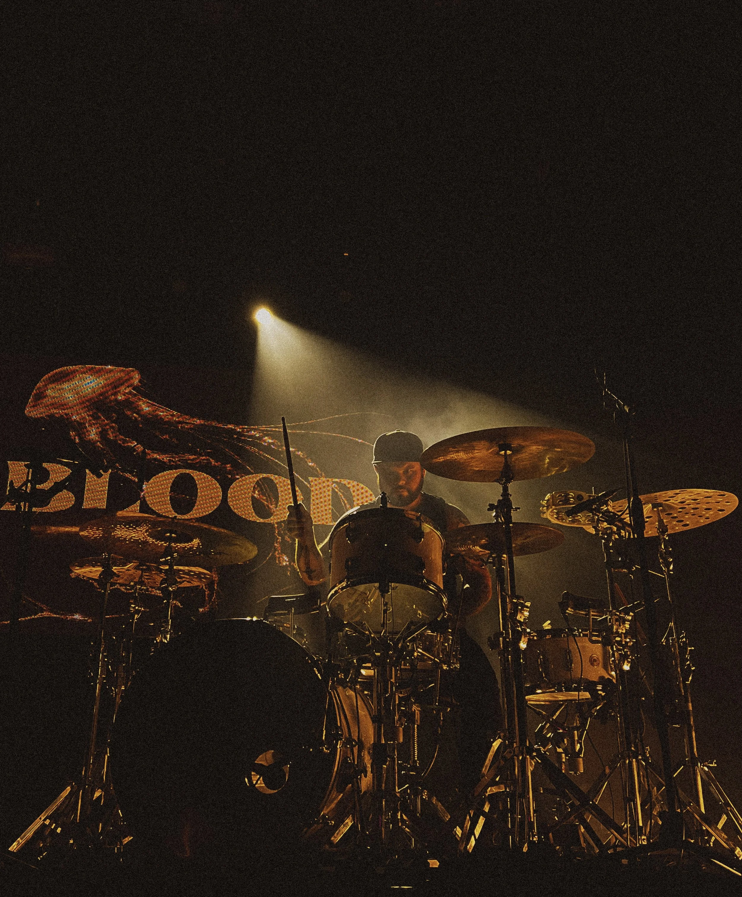 A drummer performing on stage with a dark background and a spotlight shining down, with a large backdrop that features a jellyfish and the word "BOO."