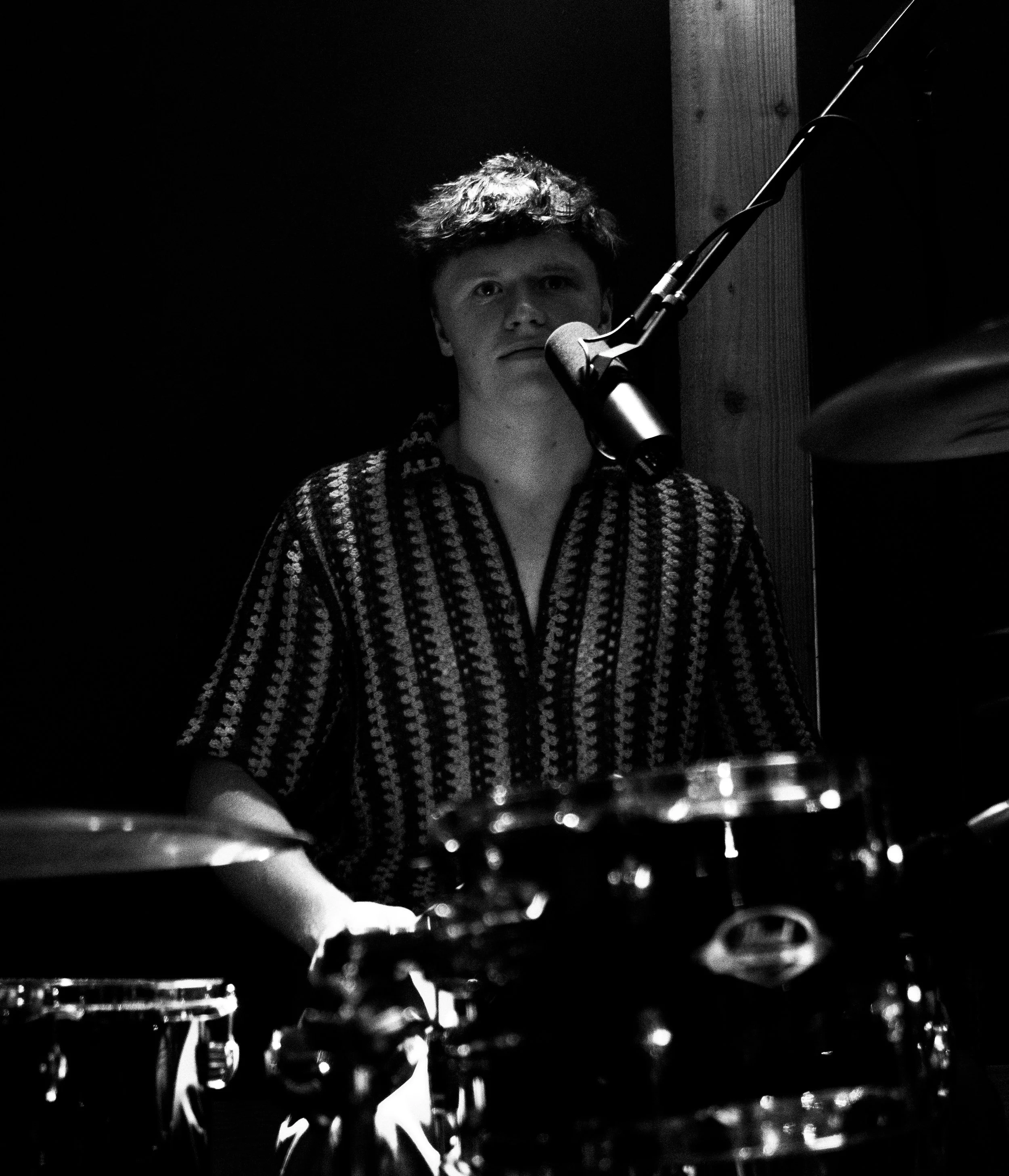A young man playing drums and singing into a microphone in a dark room, illuminated by a spotlight. Sam Beaty from Molly Gone Mad