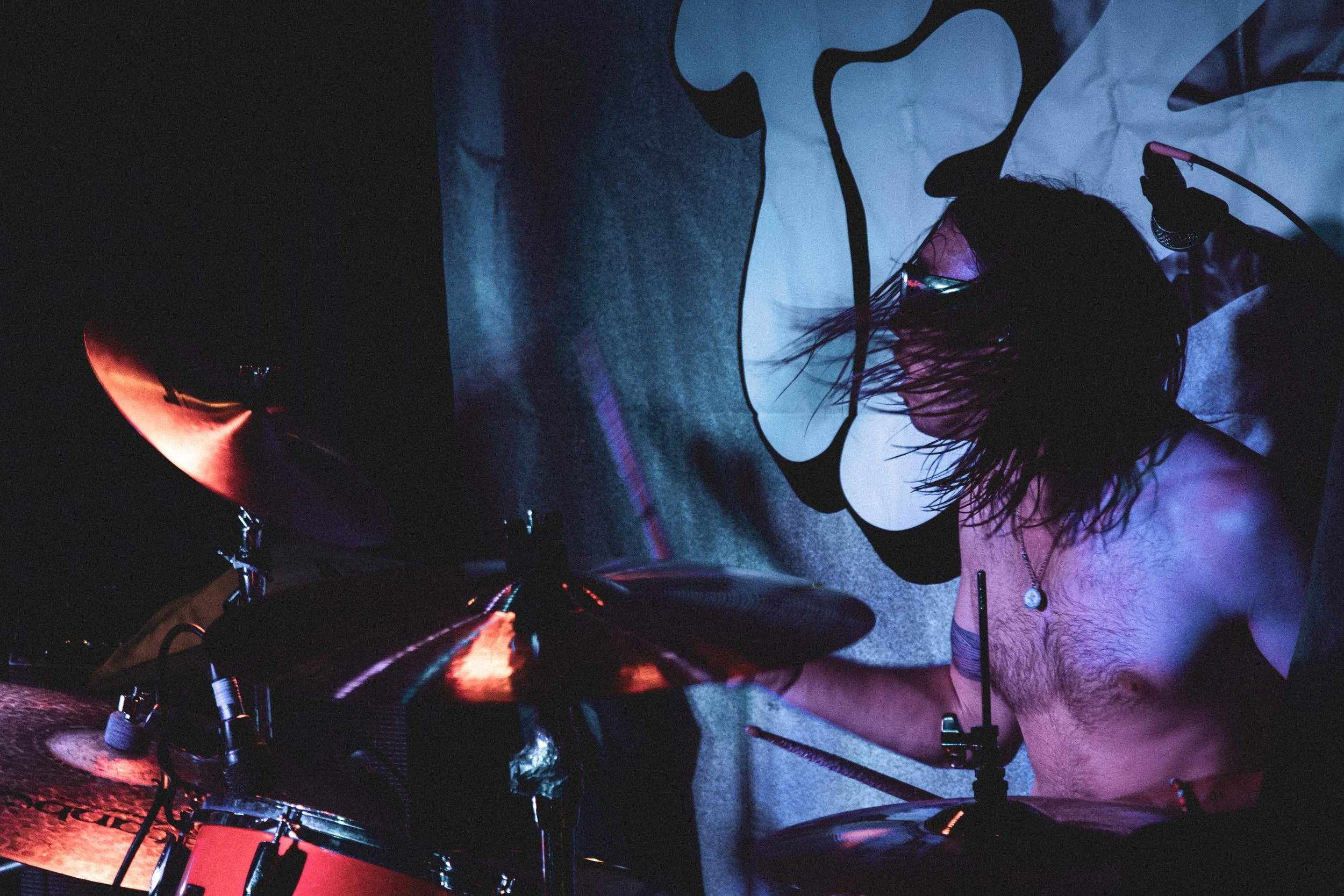 A shirtless drummer with long, dark hair playing a drum kit in a dark room with a colorful backdrop.