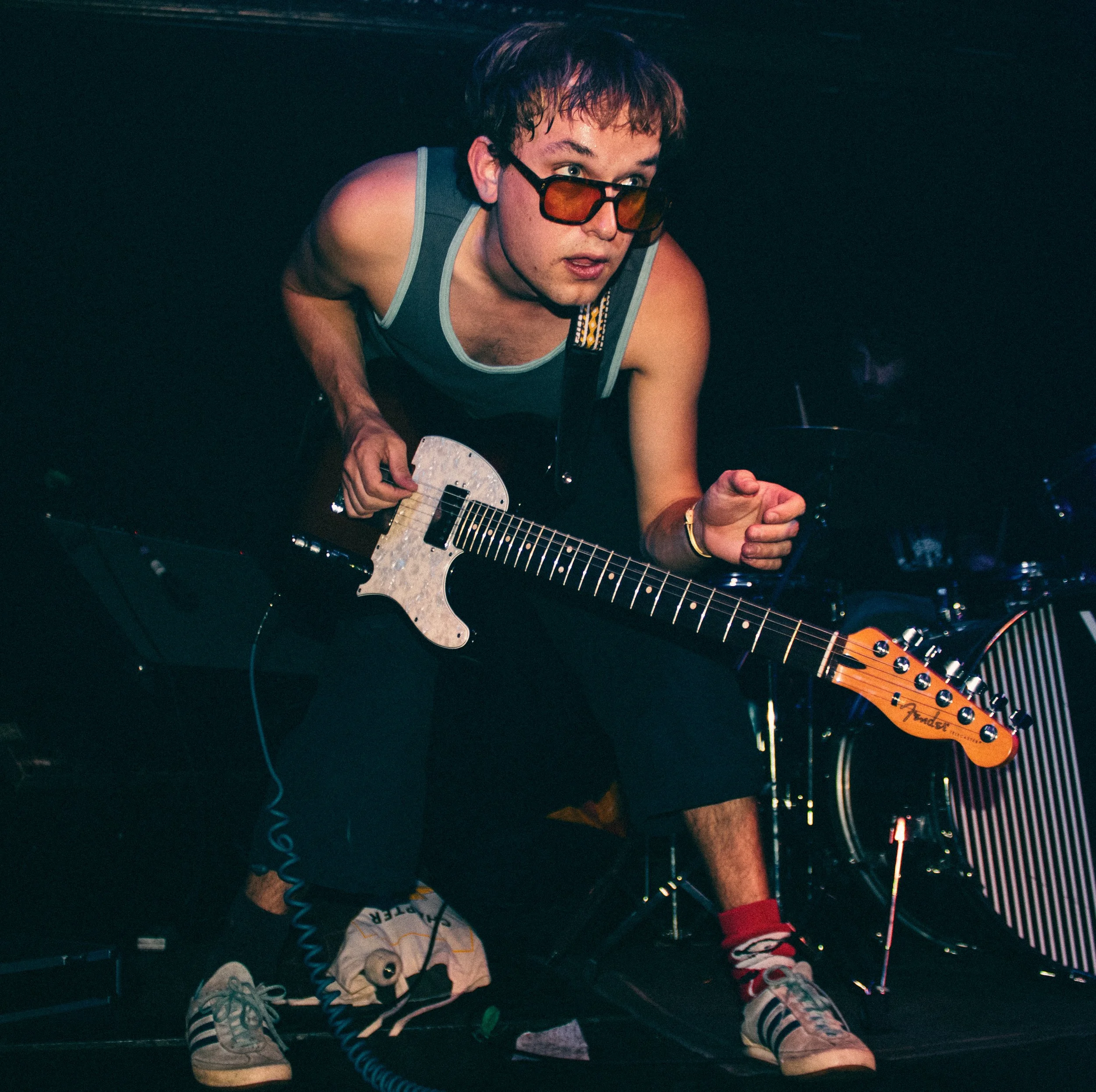 A young man with brown hair, sunglasses, sleeveless shirt, and multicolored socks plays an electric guitar on stage with a drum set in the background.