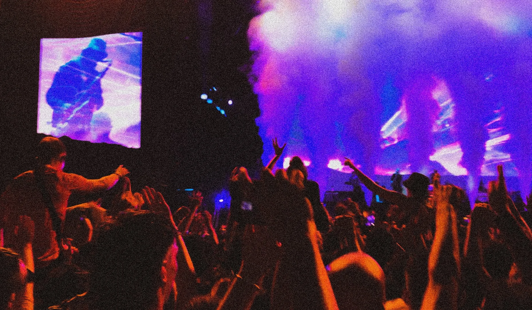 People dancing and enjoying a live music concert with colorful lights and smoke, with a large video screen in the background showing a performer. Sam Fender at TRNSMT 