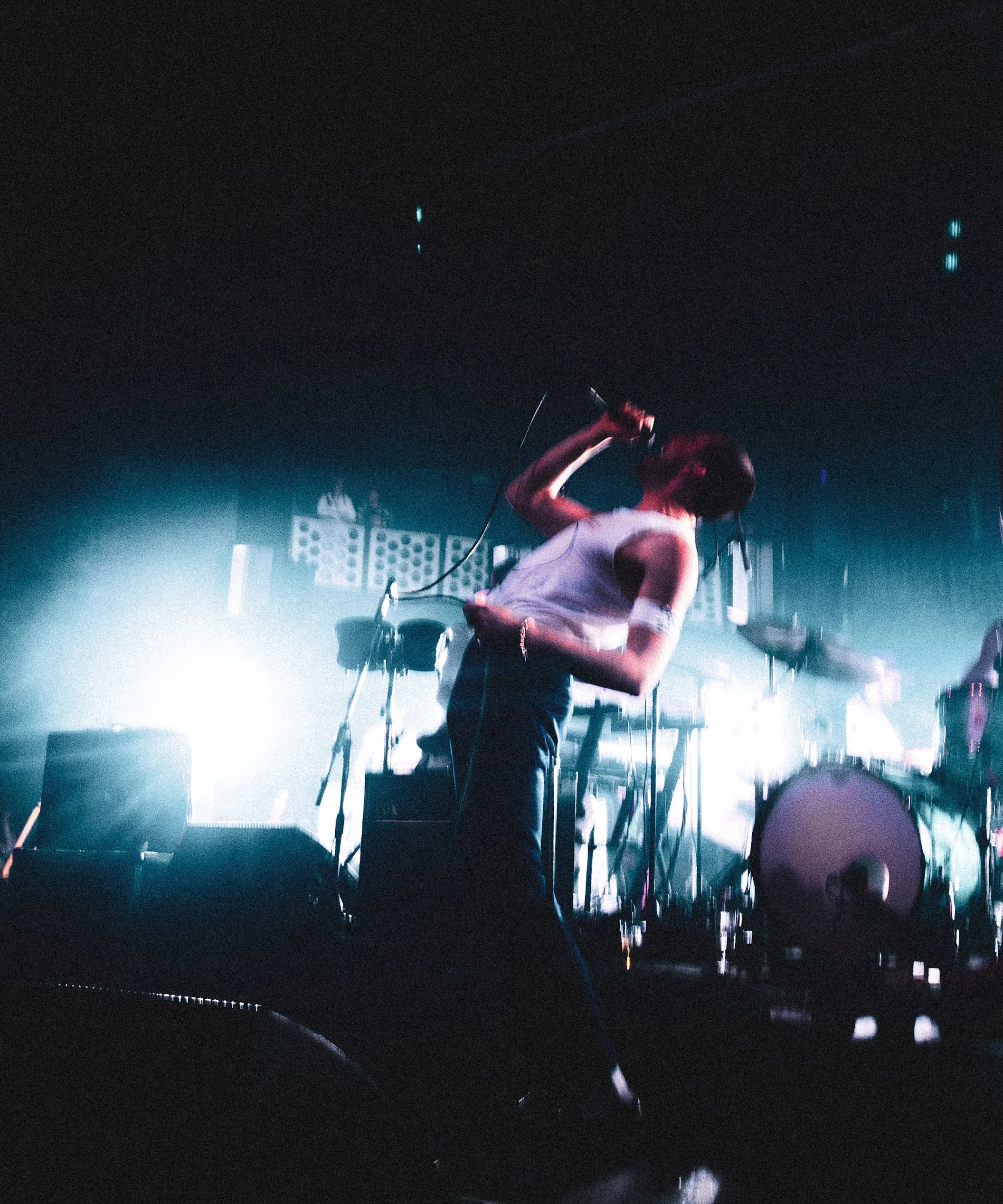 A singer performs with a microphone at a concert, illuminated by vibrant stage lights, with musical equipment and a drummer in the background. Miles Kane in the One Man Band tour, at the 02 Academy Bristol.