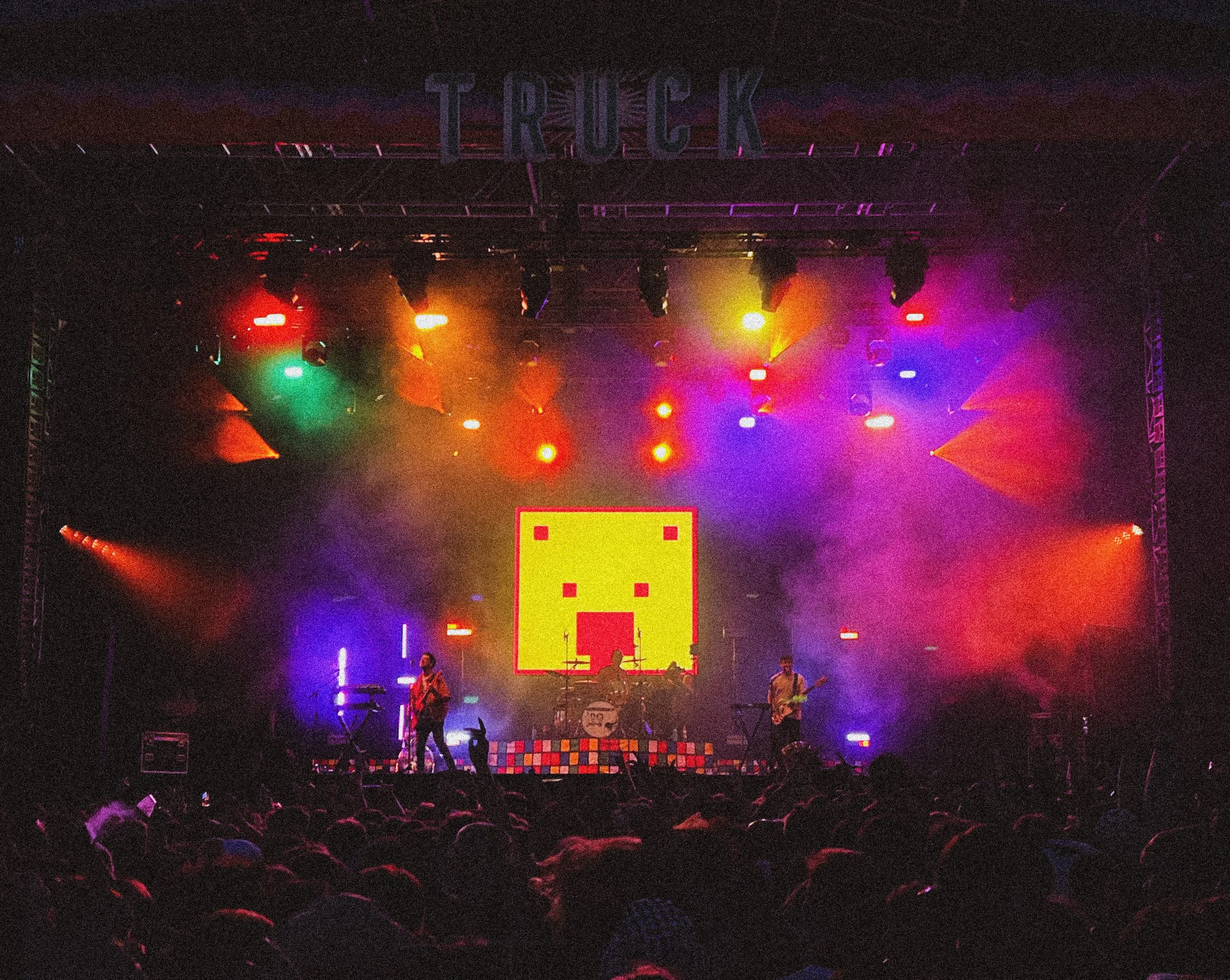 A stage with colorful lighting and a digital screen displaying a pixelated smiley face with a bow tie. A band is performing with three members visible, playing guitars and drums. The audience is in front of the stage, watching the performance.