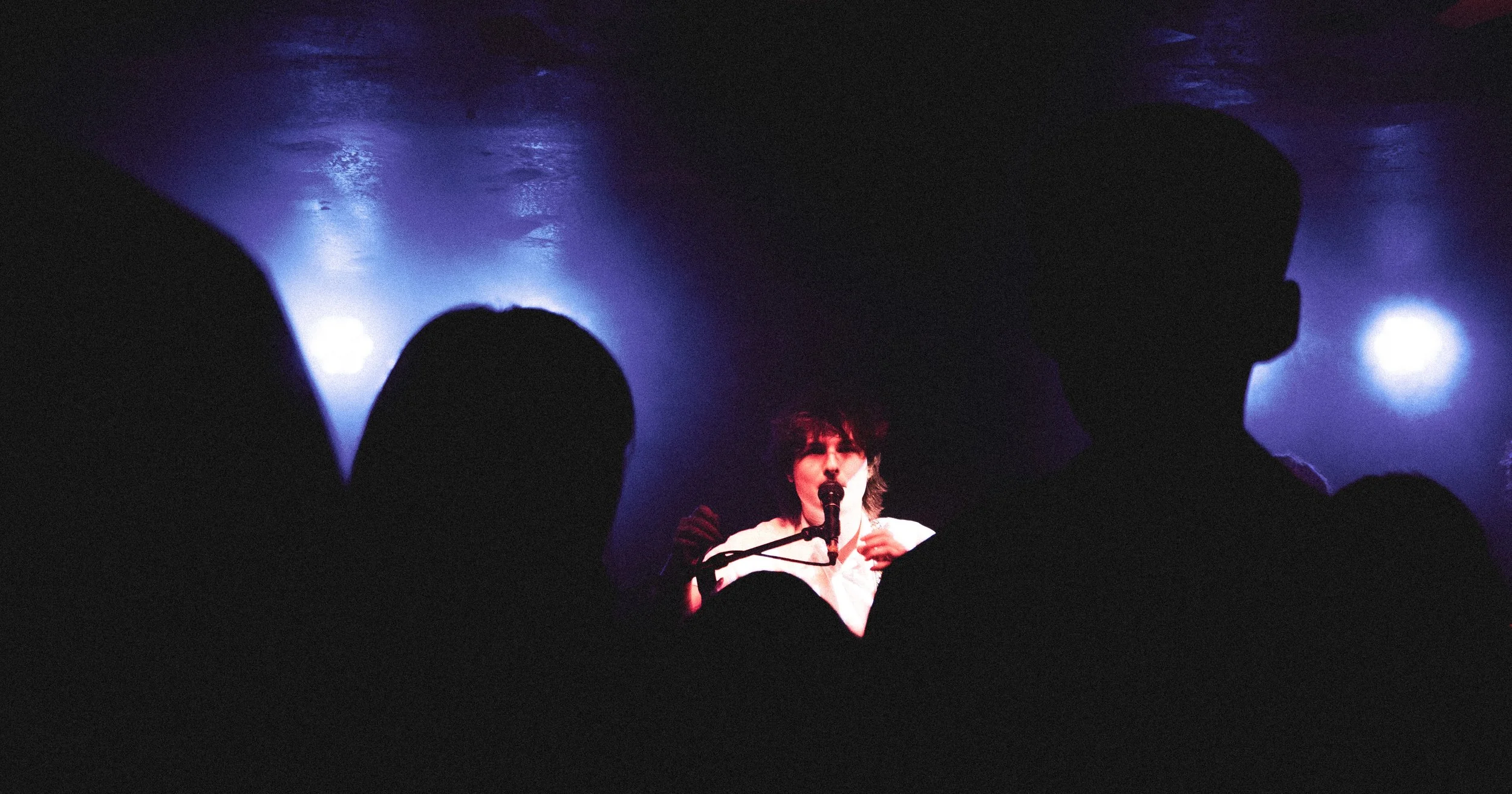A performer singing into a microphone on stage, viewed from the audience with silhouettes of people's heads in the foreground, blue and purple stage lighting.