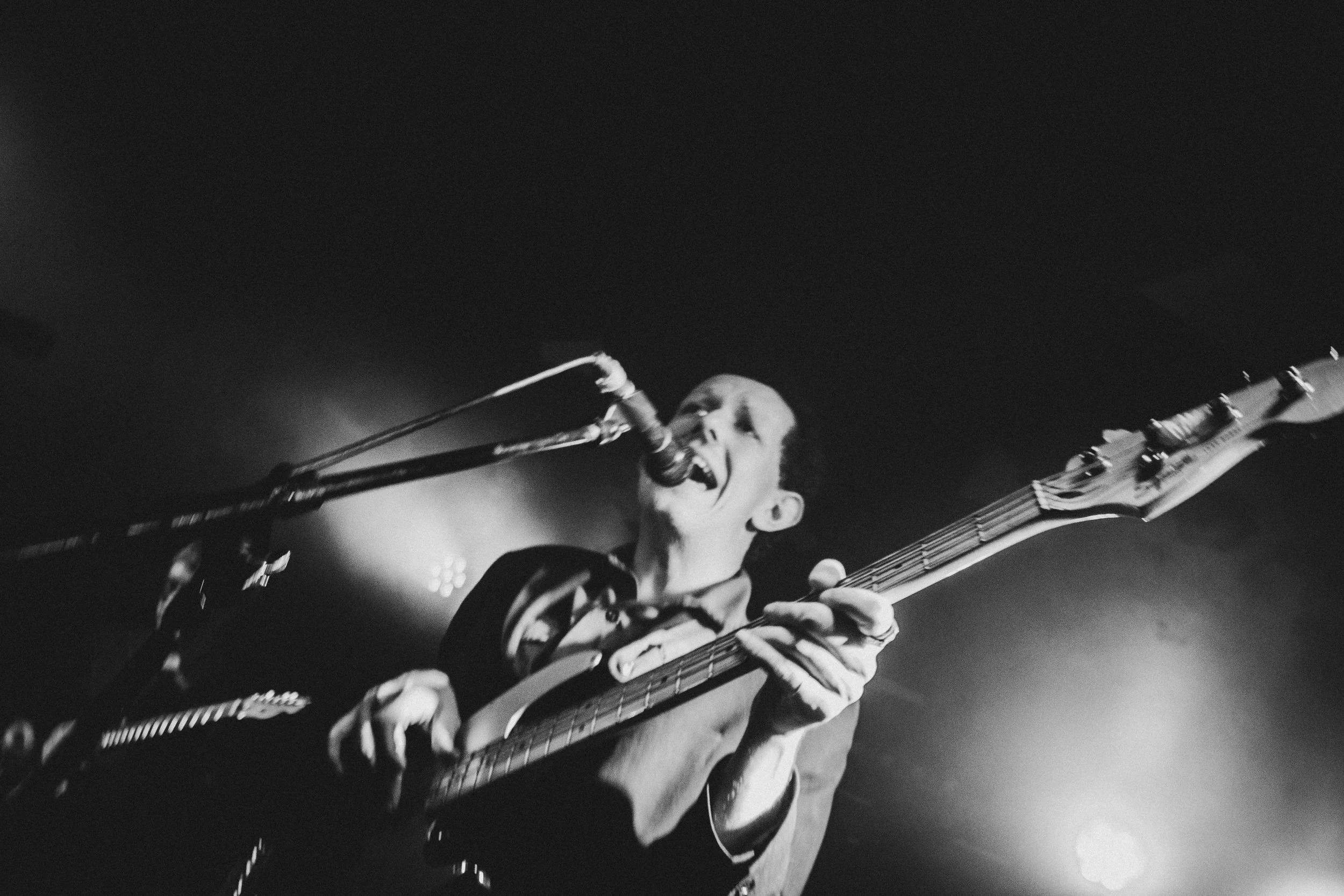 Black and white photo of a musician passionately singing into a microphone and playing an electric guitar on stage, with stage lights creating a bright glow in the background.