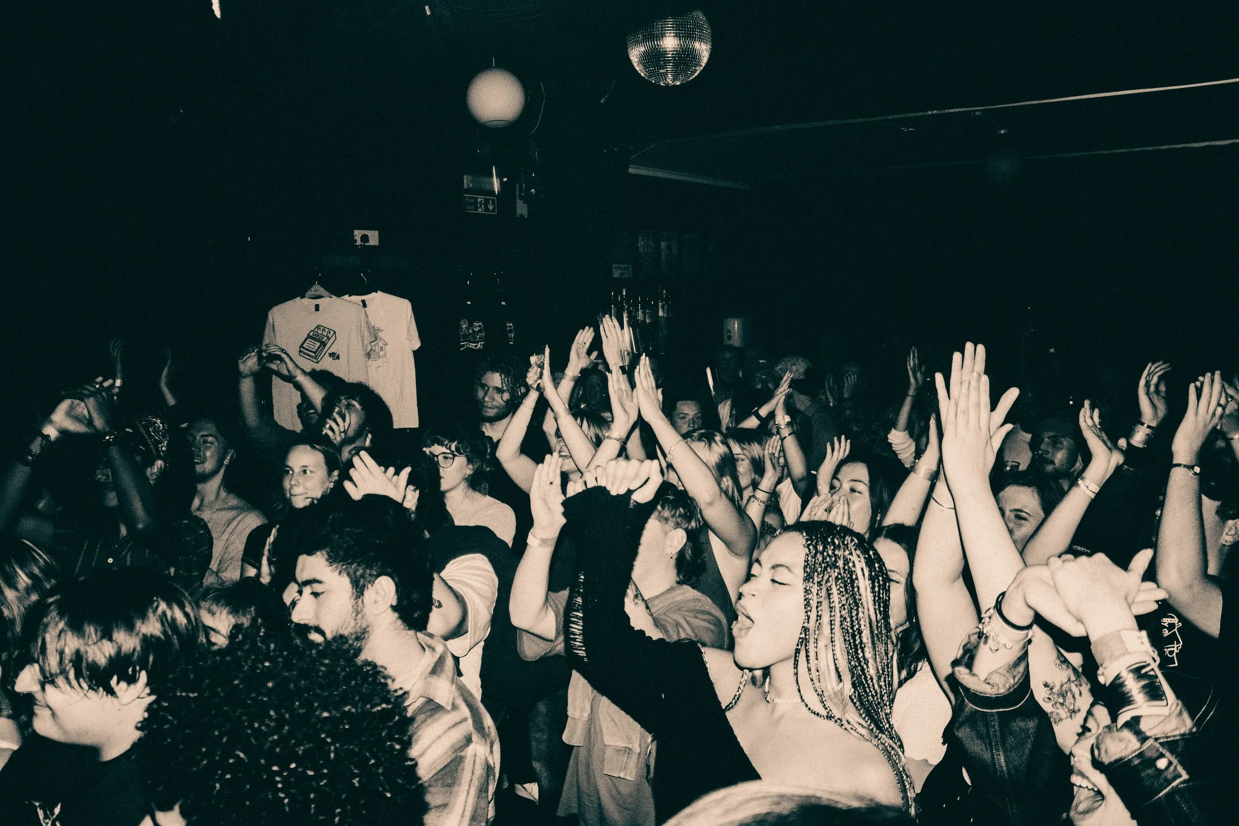 Crowd of people at a dance party with their hands raised, dancing and enjoying the music, in a dark room with disco balls overhead.