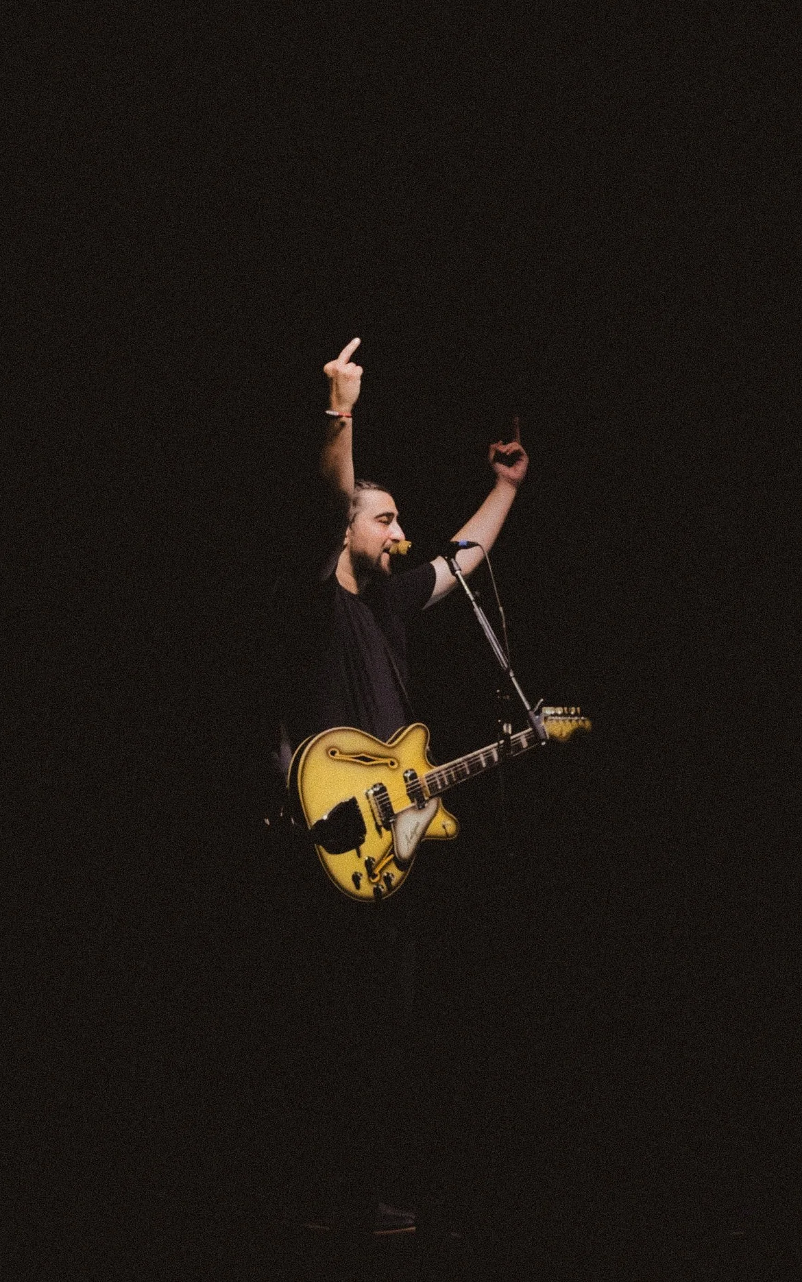 A musician on stage playing an electric guitar with a yellow body, raising both arms with middle fingers extended into the air, surrounded by darkness. Noah Kahan, Cardiff