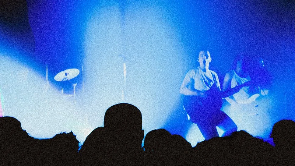 A live music concert with a singer and guitarist on stage, illuminated by blue lighting, audience silhouettes in foreground, and drum set in background.