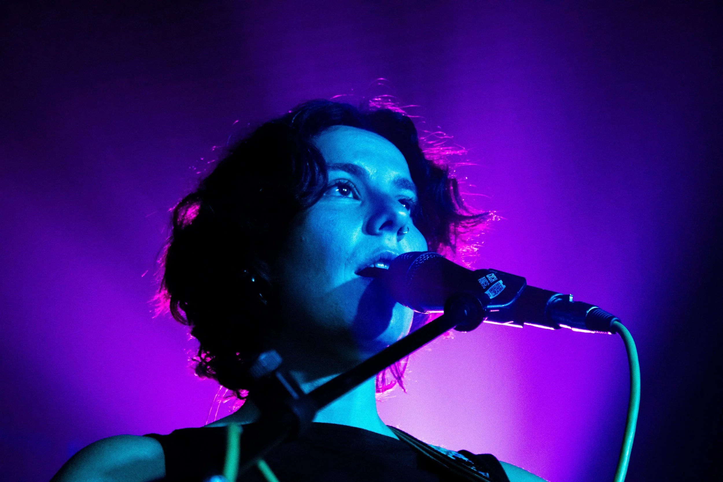 A woman with dark curly hair singing into a microphone, illuminated by purple and blue stage lighting.