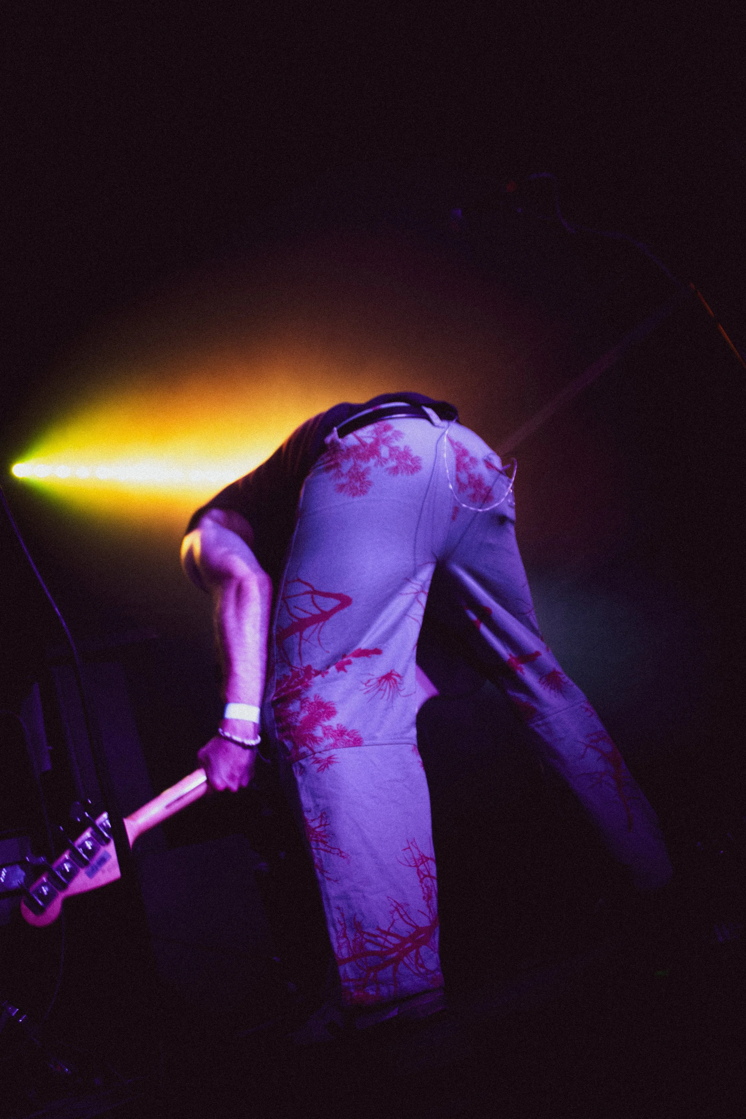 A person on stage with colorful purple, yellow, and red lighting, wearing light-colored pants with a red tree pattern, holding a guitar, facing away from the camera.