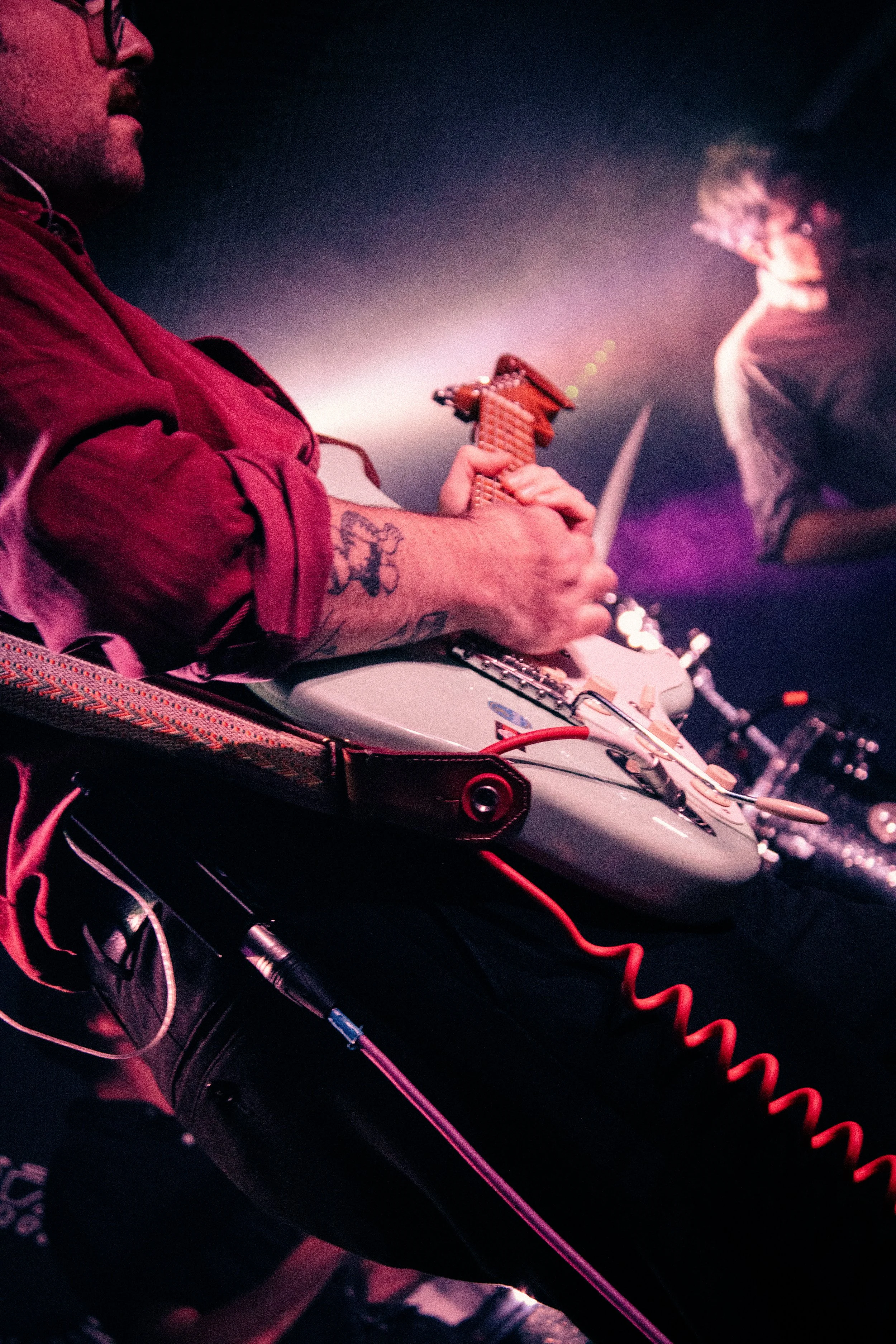 A musician in a red shirt plays an electric guitar on stage, with a tattoo on his forearm and a woman in the background with curly hair and a mask on, illuminated by purple stage lights.