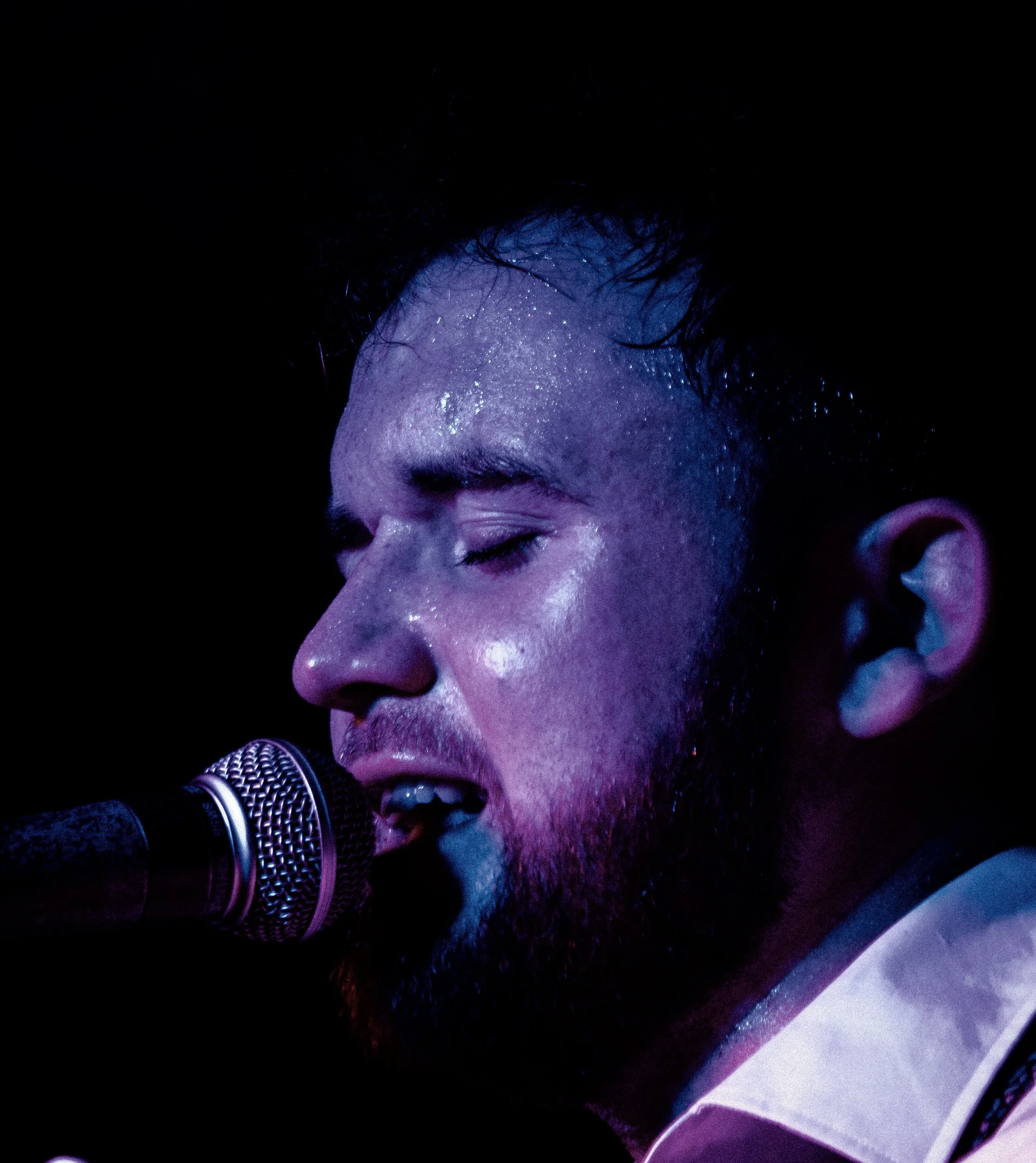 Close-up of a male singer with closed eyes singing into a microphone, illuminated by purple stage lighting, with beads of sweat on his face.
